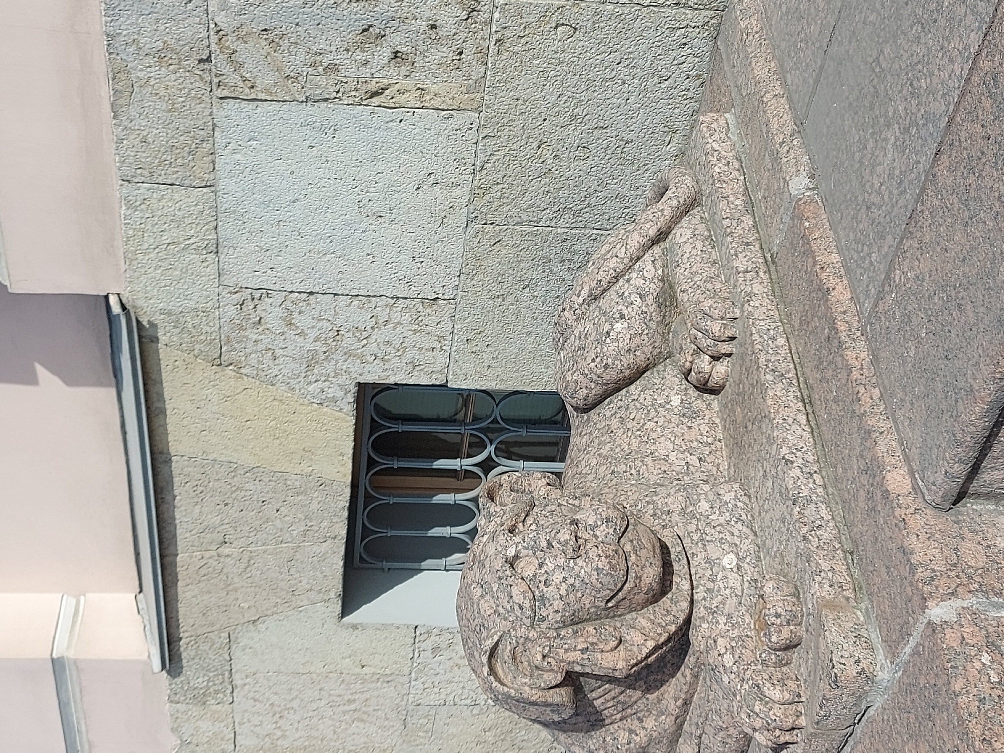 A close-up outdoor shot presents a granite sphinx sculpture positioned adjacent to a building facade. The building features light-colored stone blocks and a window with a metal grill. The sphinx, crafted from a reddish-pink speckled granite, is intricately detailed, showing the sphinx's head, paws, and a portion of a stone book or tablet it is holding. The composition is framed from a low angle, emphasizing the statue's size and detail in relation to the building's architecture. The scene is likely set in Saint Petersburg, Russia, as these sculptures are common there. The overall ambiance suggests a historical or cultural site.