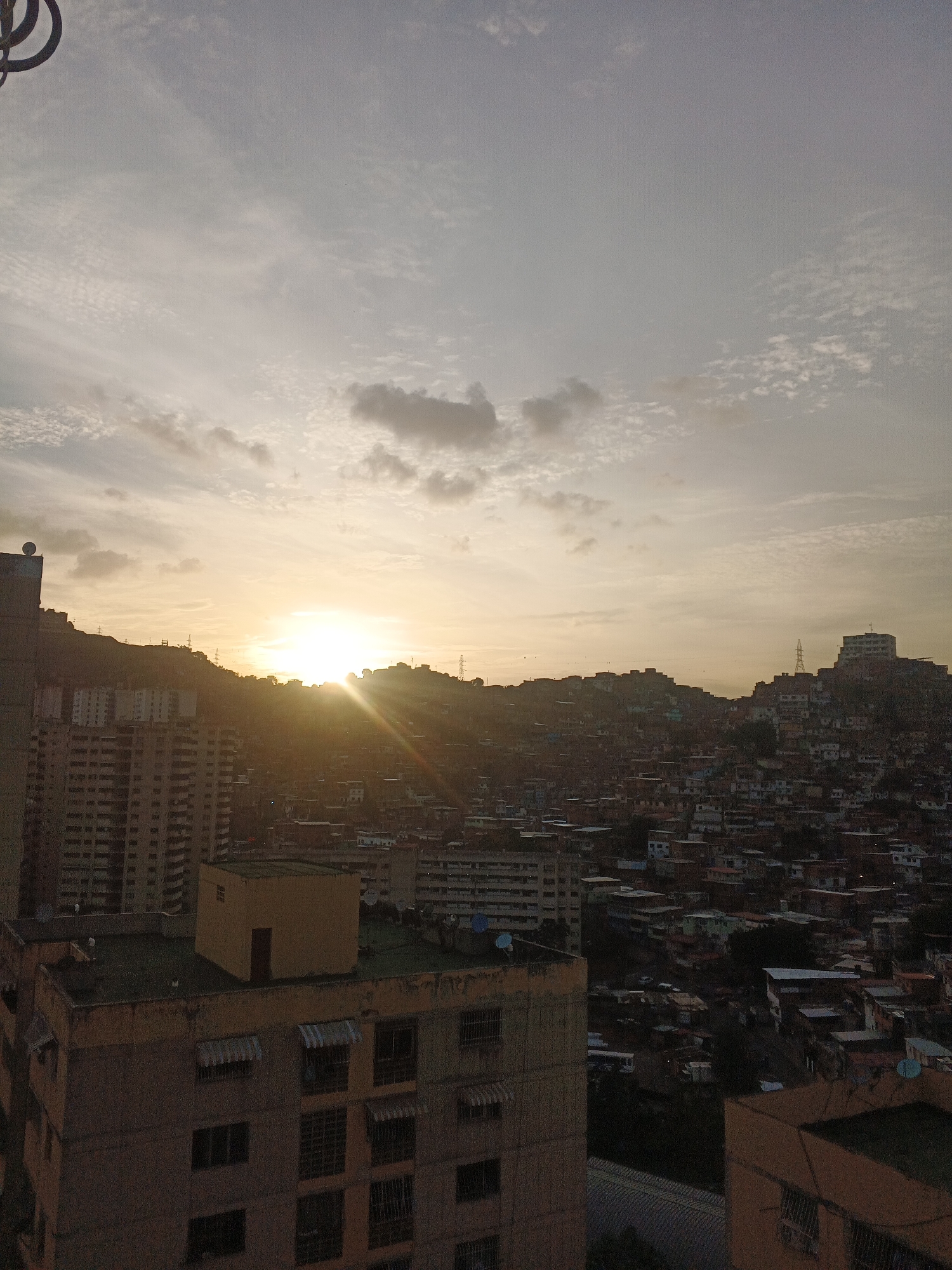 The image shows a panoramic view of Caracas, Venezuela, at sunset. The sun is setting behind a mountain range in the background, casting a golden light over the city. In the foreground, there are several apartment buildings, and beyond that, a densely populated area with a mix of residential and commercial buildings. The sky is filled with clouds, and the overall scene is hazy due to the atmospheric conditions.