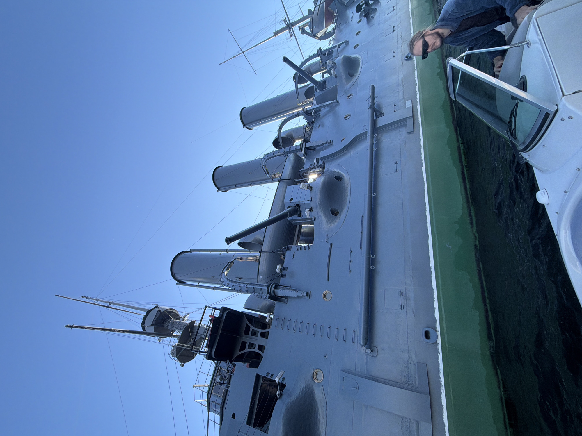 A low-angle, wide shot shows a man in sunglasses and a blue jacket sitting in a white boat, looking to his right. The boat is on dark water. Behind the boat is the side of a large, gray ship with a green waterline. The ship has multiple smokestacks, gun turrets, and masts, suggesting it is an older warship. The sky above is clear and blue. The scene is likely a tourist attraction in an unknown city.