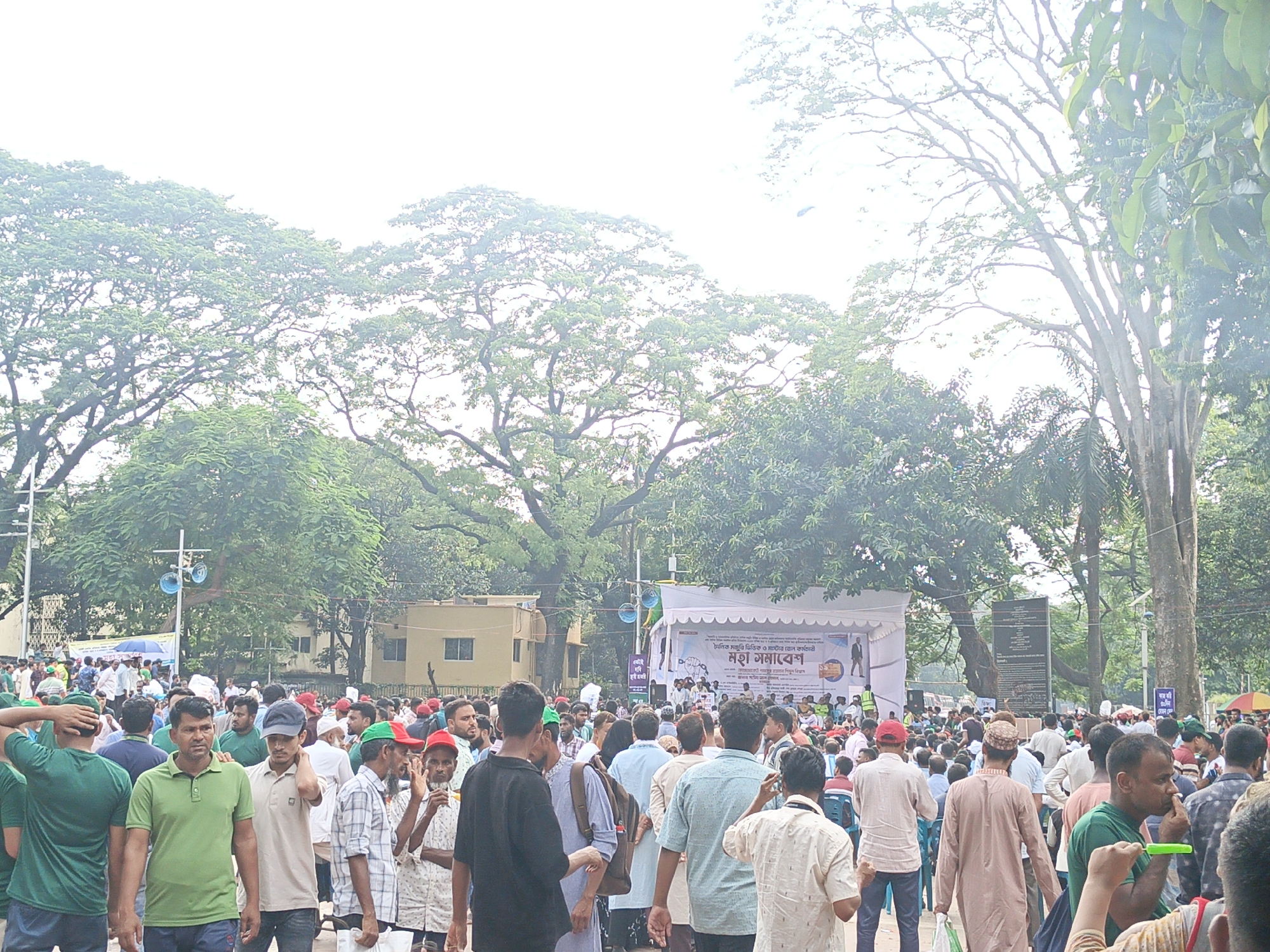 The image depicts a large outdoor gathering of people in an urban setting. A stage with a banner displaying Bengali text and graphics is set up in the background. Numerous individuals, predominantly men of various ages, are present, some wearing casual attire, while others are in traditional clothing. Many are standing, some are walking, and a few appear to be engaged in conversation. The environment is characterized by lush green trees providing shade, and the presence of buildings and poles suggests a developed area. The sky is overcast, indicating diffused lighting conditions. The overall scene conveys a sense of a public event or rally.