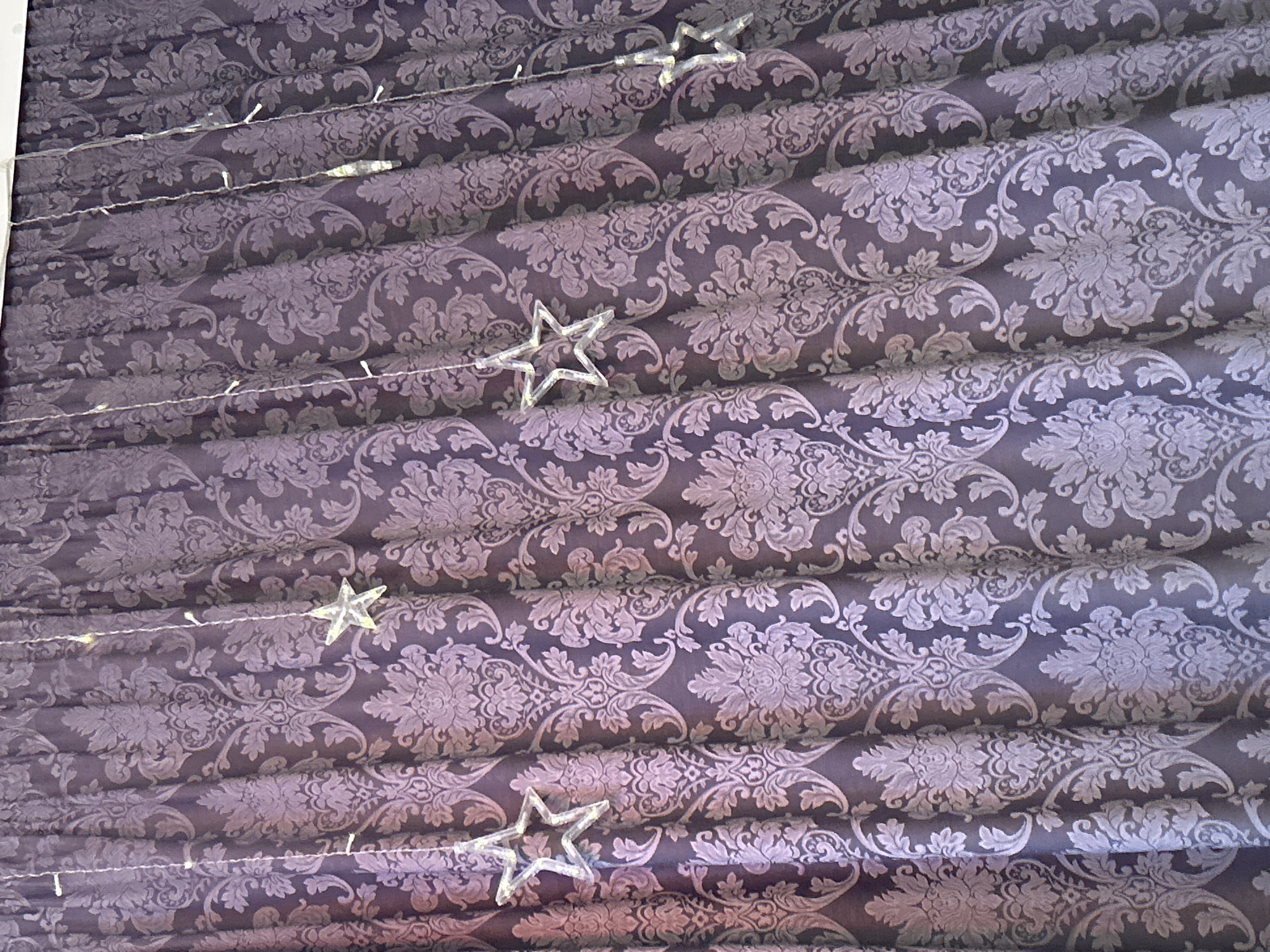 The image shows a folded curtain with a dark purple, floral pattern. Decorative, star-shaped lights are interspersed across the curtain folds. The location is Chaikovsky, Russia. No people are visible.