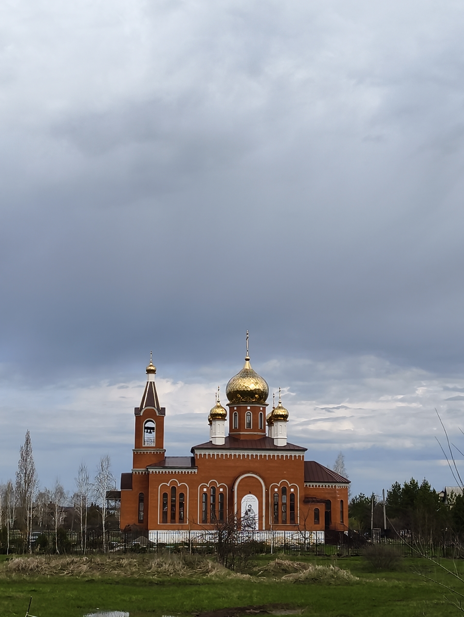 The image features a brick Orthodox church located in Balashov, Russia. The church has multiple golden domes and a bell tower. The sky is overcast with dark clouds. The foreground consists of a grassy area. Trees are present in the background.