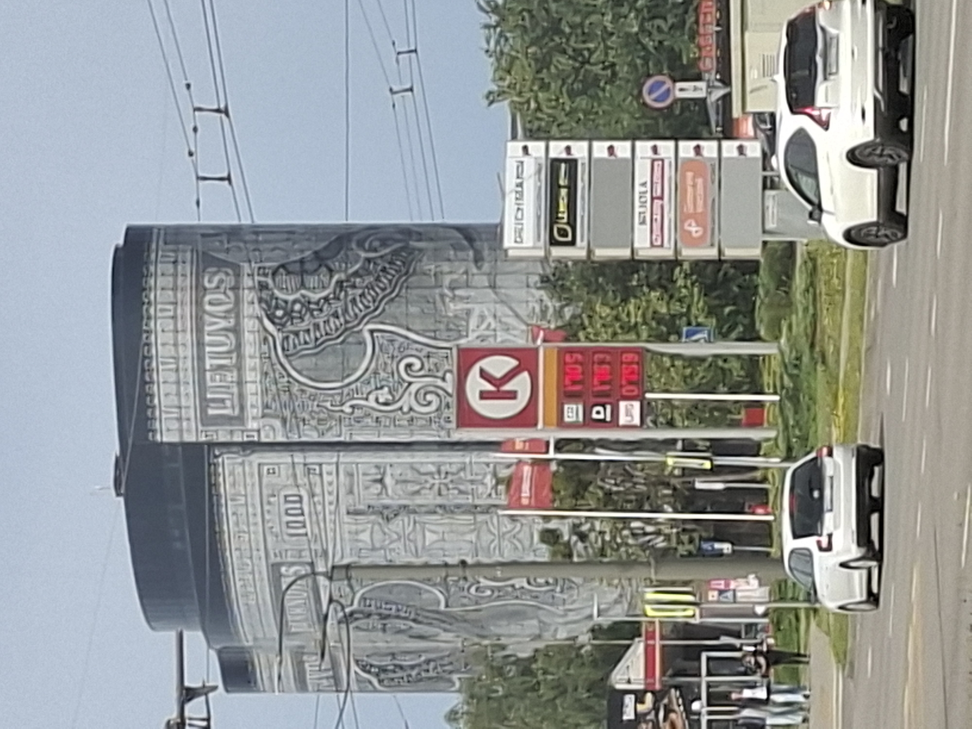 The image captures a street scene in Kaunas, Lithuania, featuring a large cylindrical building with a mural on its facade. The mural appears to depict intricate, possibly abstract or historical, designs and includes text that reads "LIETUVOS" and "1000". In the foreground, a white Skoda Octavia hatchback is driving on a paved road. To the right of the Skoda, a white Honda CR-V is also driving on the road.
Adjacent to the cylindrical building is a Circle K gas station, indicated by the red "K" logo and illuminated price displays showing fuel costs such as "1705", "159", and "0759". Several signs are stacked vertically next to the gas station, displaying various brand names or services, including "DRICHEMAC", "LIUCIN", and "SUJOLA". Red and white flags are visible behind the gas price display.
The environment suggests an urban setting with trees and overhead power lines. A few pedestrians are walking on a sidewalk in the background, near what appears to be a commercial establishment with red accents. The sky is overcast.