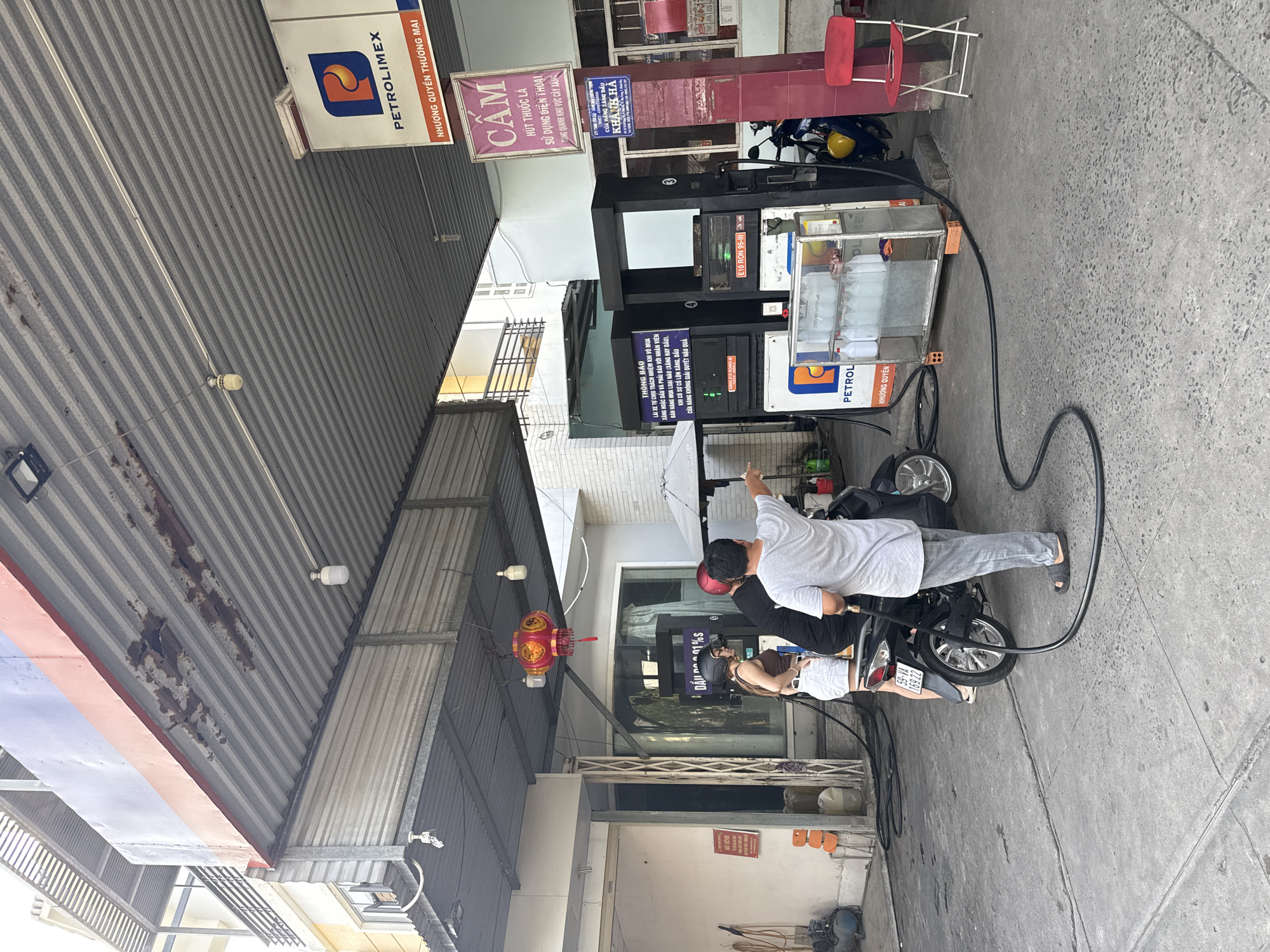 At a petrol station in Bac Nha Trang Ward, Vietnam, a man in a grey t-shirt and grey pants is standing next to a motorcycle, pointing to the right while holding a black fuel hose. He is facing a woman wearing a brown tank top and white shorts, who is also standing next to the motorcycle and wearing a helmet. A person in a black shirt is behind the woman, also wearing a helmet. The motorcycle is black with a license plate reading "59-V4 169.22". The petrol station has a corrugated metal roof with visible rust and a PETROLIMEX logo. Red and gold lanterns hang from the ceiling. A sign with Vietnamese text that translates to "No smoking. Do not use mobile phones. Do not honk." is visible. Another sign in Vietnamese advises about fuel purchases. A red folding chair is positioned near a red support column. The ground is paved with concrete.