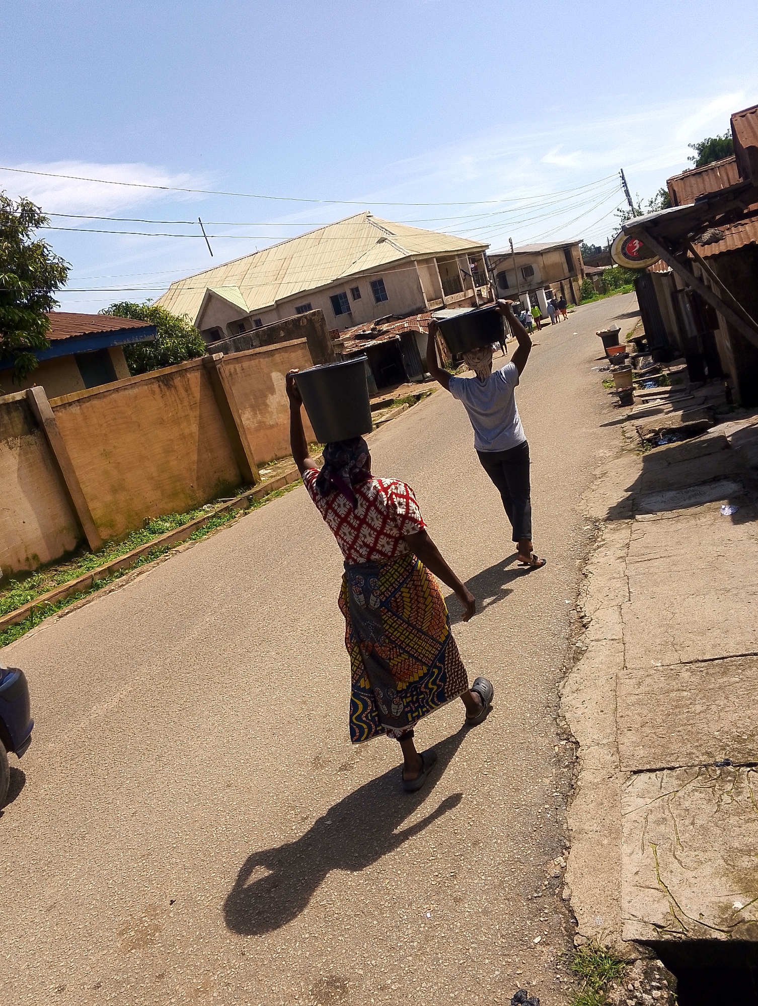 Two people are walking on a paved road under a bright, sunny sky, suggesting it is daytime. The person in the foreground, a woman, is walking away from the camera and carrying a large black bucket on her head. She is wearing a colorful, patterned dress. The second person, who appears to be a man, is walking in front of her, also carrying a bucket on his head. He is wearing a light-colored t-shirt and dark pants. The road is lined with buildings on either side. On the left, there is a long, tan-colored wall with some green foliage growing alongside it. Beyond the wall, a building with a light-colored roof and a blue trim is visible. On the right side of the road, there are several structures with corrugated metal roofs, some of which appear to be small shops or stalls. The overall scene depicts daily life, likely in a community where carrying water in buckets is common. There is no visible text in the image.