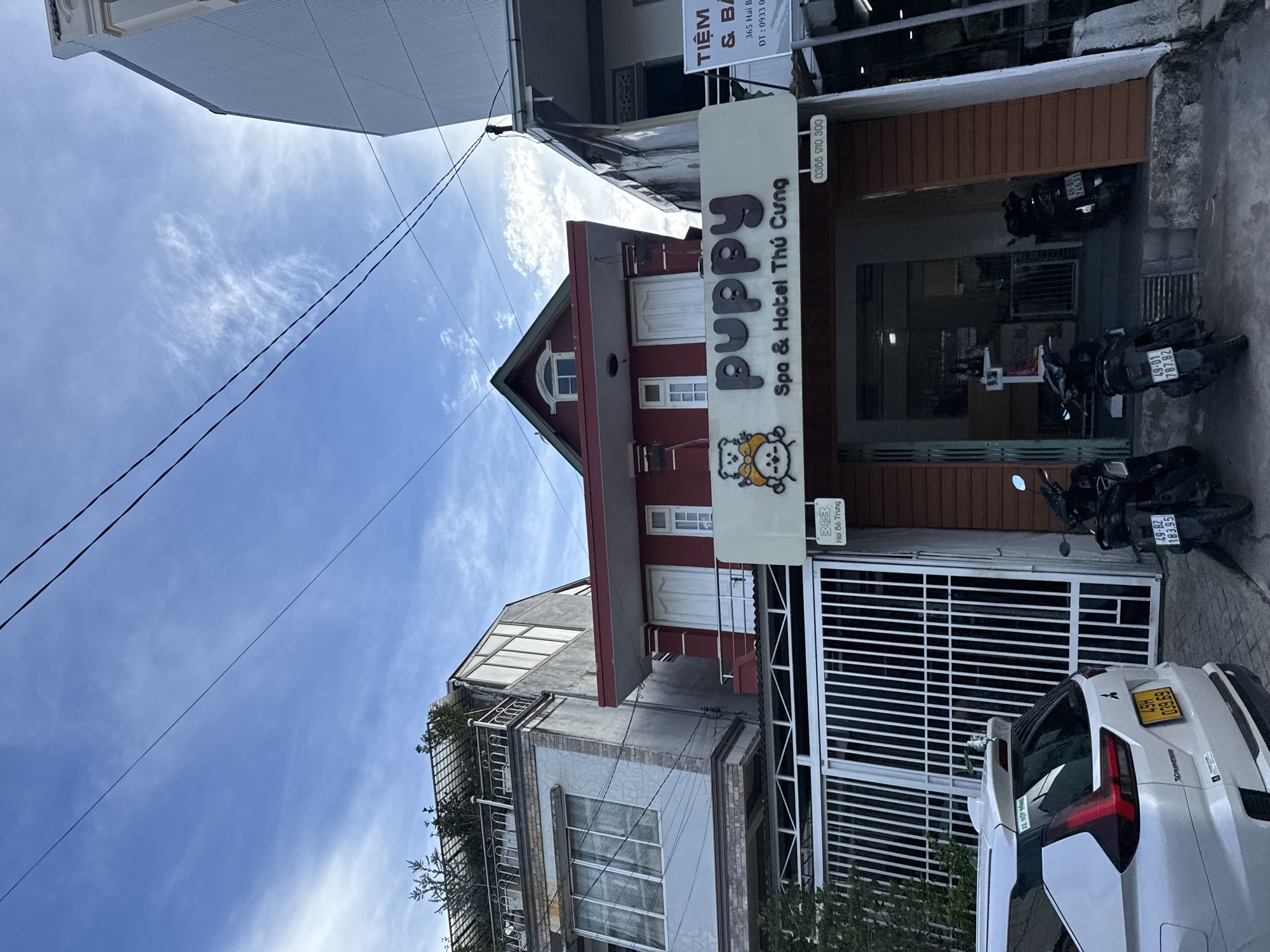 The image captures a street view in Da Lat, Vietnam, featuring a red building with a sign for "Puppy Spa & Hotel Thú Cưng." The building's facade is adorned with white shutters and a small attic window. To the left of the red building, a gray and white structure with a balcony and plants on its roof is visible, and to the right, a white building with a sign that partially reads "TIỆM & BA..." and a phone number is present. In front of the red building, a white gate stands, and a white SUV with a Vietnamese license plate is parked on the left. Several motorcycles are parked near the entrance of the spa and hotel. The sky above is blue with scattered clouds and power lines crisscrossing the frame.