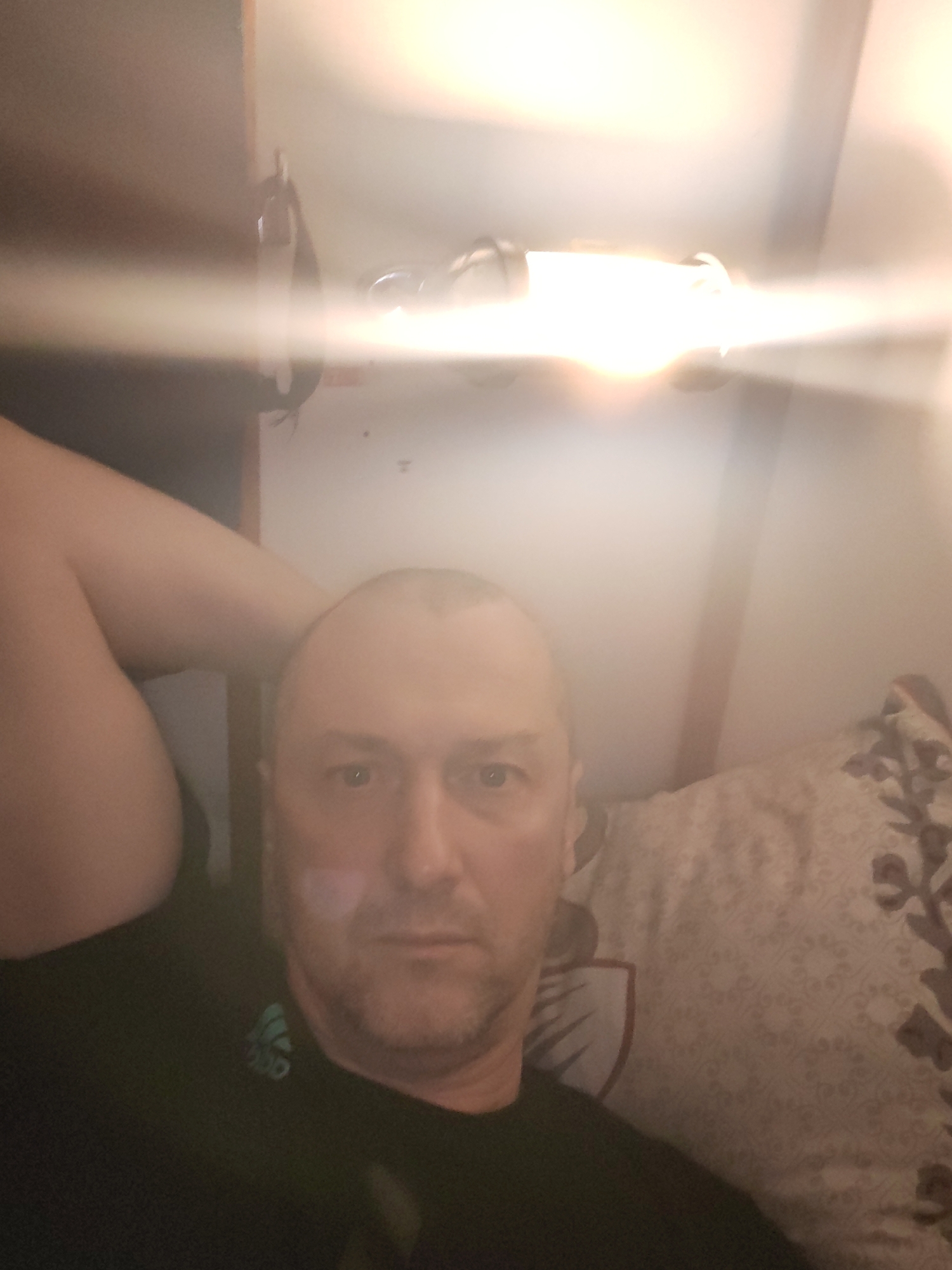 The image is a close-up, slightly blurry selfie of a man taken indoors. He appears to be reclining, with his right arm bent and resting behind his head or shoulder. The man has short or receding light-colored hair and short stubble or a beard. He looks directly at the camera with a neutral expression.
He is wearing a black t-shirt with a light green or teal logo visible on the left side of his chest, which resembles a stylized sports brand emblem, possibly similar to the Adidas trefoil.
A very bright artificial light source, likely a bare bulb or light fixture, is prominent in the upper right portion of the frame. This light creates a strong horizontal lens flare that spans across the entire image, obscuring some details of the man's face and most of the background.
Behind the man, to his right, is a pillow with a light-colored, possibly beige or cream, fabric adorned with a darker brown floral or geometric pattern. The background beyond is indistinct due to the intense light and blur, but appears to be a wall with a darker element or shadow on the far left. The overwhelming brightness from the artificial light suggests the photo was taken in a dimly lit environment, possibly at night or in the evening. There is no other discernable text or specific location cues beyond the indoor setting suggested by the pillow.