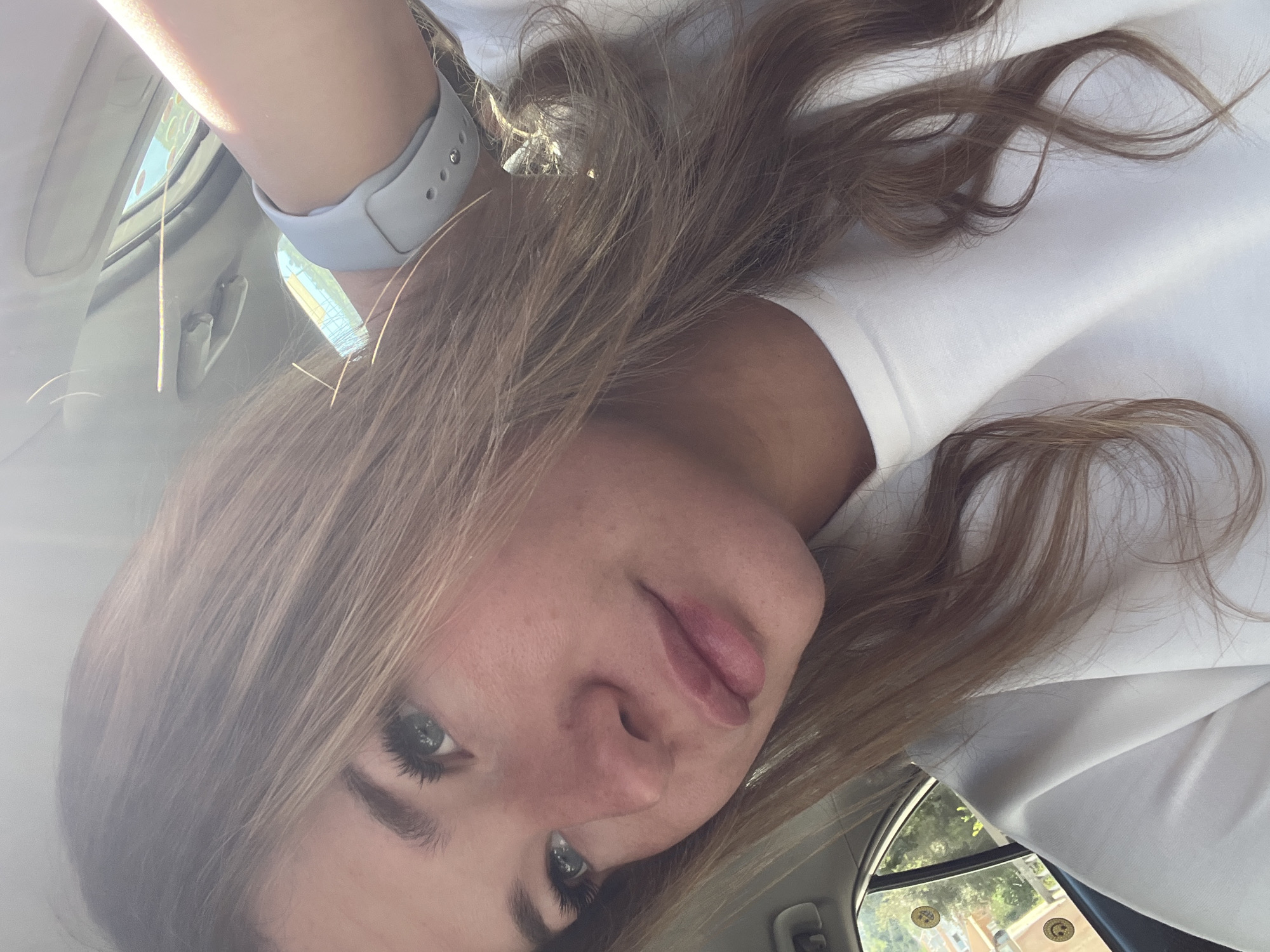 A medium close-up shows a woman with shoulder-length brown hair, fair skin, and light eyes. She is wearing a white shirt and a gray smartwatch. The background reveals the interior of a car, and through the window, a glimpse of buildings and trees is visible, suggesting an urban environment. The image is angled, possibly taken from a slightly elevated position or with the camera tilted. The location cannot be confidently confirmed as Budva, Montenegro, based on the visual information.