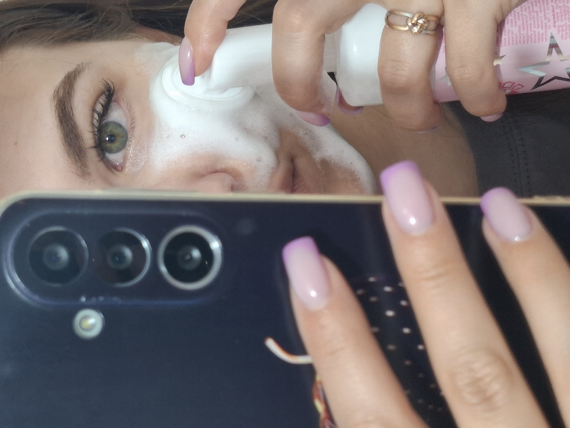 A selfie-style image features a person applying white foam onto their face. The person's left eye is visible, and it is green. The person has manicured nails with a light pink base and a purple tip. The person is holding a pink bottle with white pump. The person wears a gold ring. The phone used to take the image is visible in the foreground and has three camera lenses and a flash. The image does not show any locations to be able to establish the place of the photo.