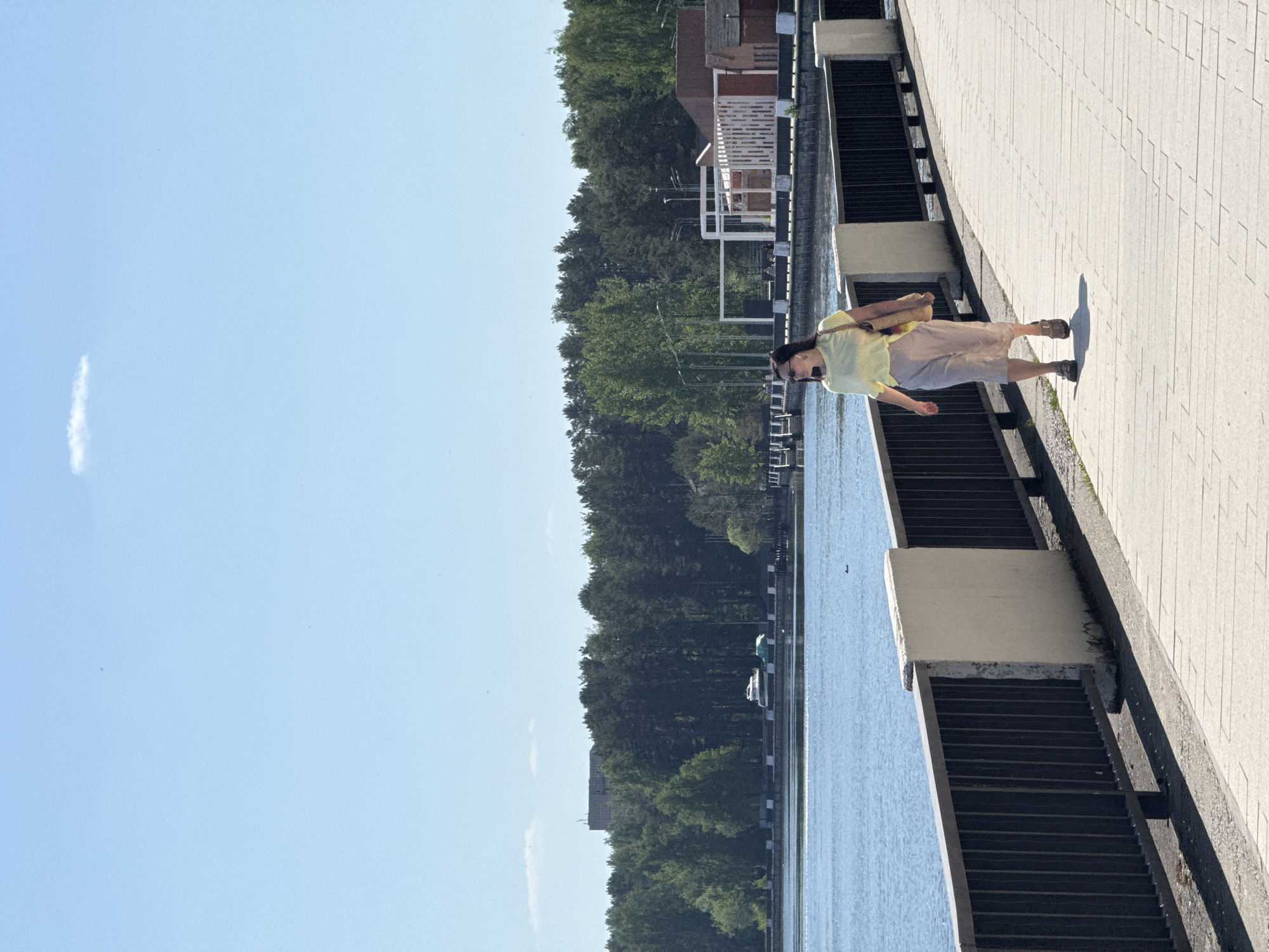 A woman walks along a light gray paver walkway alongside a body of water under a clear blue sky with a wisp of cloud. The walkway is bordered on one side by a dark gray railing with concrete pillars and on the other by a line of trees. Beyond the trees are buildings. The woman is wearing a light yellow shirt, tan shorts, and sandals. She is carrying a bag over her shoulder. The location is Unknown, Unknown.