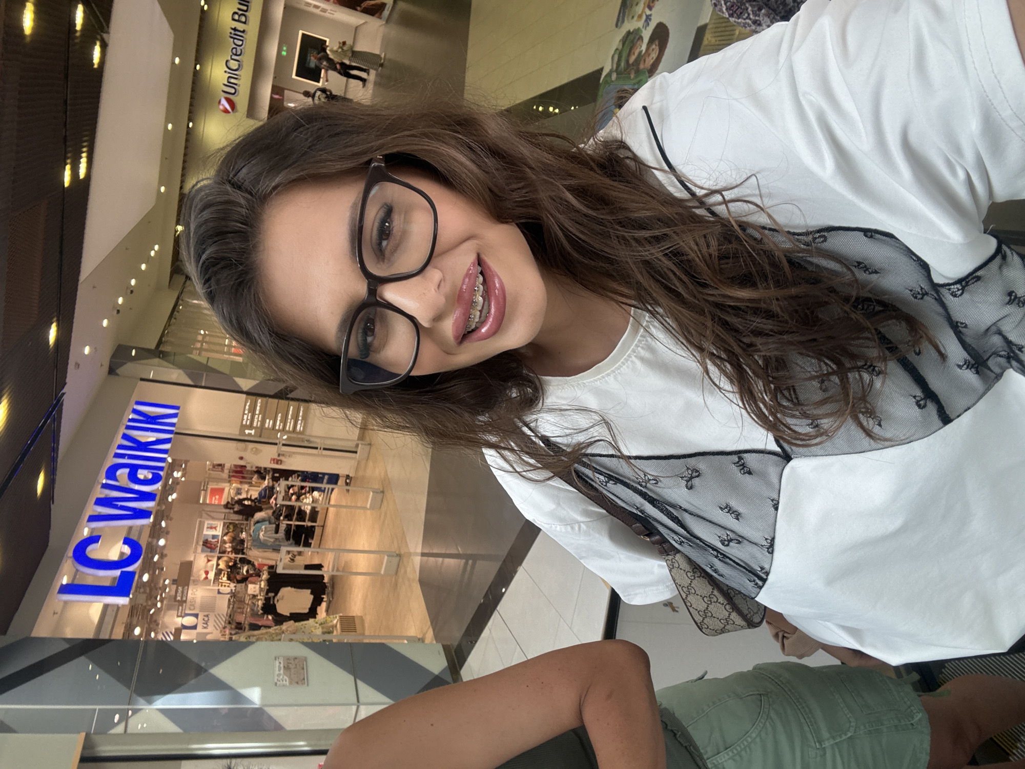 The image shows a woman with brown hair and glasses, wearing a white t-shirt with black lace detailing, and braces on her teeth. She is taking a selfie in what appears to be a shopping mall. In the background, the "LC Waikiki" store is visible, which is a clothing retailer. Also visible in the background is a "UniCredit Bulbank" sign. LC Waikiki and UniCredit Bulbank are both present in the Grand Mall in Plovdiv, Bulgaria, making it the likely location. The woman is holding a handbag with a patterned strap. Another person, partially visible on the left, is wearing a green top and shorts.