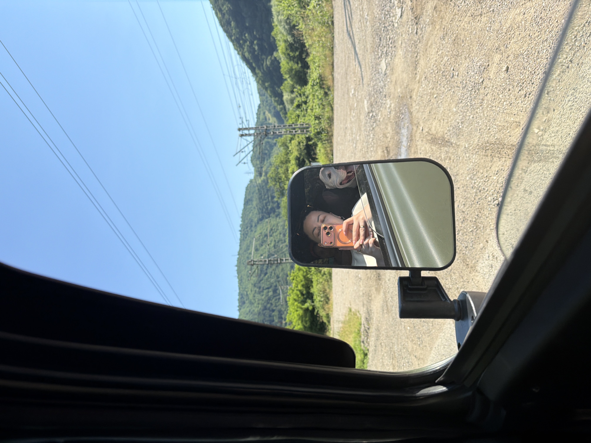 A reflection in a side-view mirror shows a young woman holding a phone in her right hand, taking a selfie. She is wearing a white top and has dark hair. Next to her, a white dog with its mouth open and tongue out is visible. The background in the mirror is a green, tree-covered mountainside with a clear blue sky above. Power lines and a utility pole are visible in the distance. The vehicle appears to be on a gravel road. The image is taken from inside the vehicle, looking out towards the side mirror. The approximate location is Krasnoye, Russia.