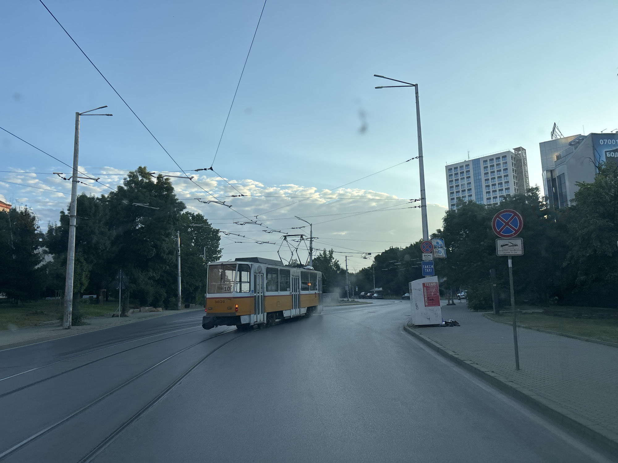 This image depicts a street scene in Sofia, Bulgaria, likely in the summer or early autumn due to the clear sky and lush green trees. The main subject is a yellow and white tram, number 3030, on tram tracks on a wide, paved road. The tram is in motion, moving away from the viewer and towards the left side of the frame. A faint mist or steam is visible behind the tram, possibly from its braking system or from the road surface.
The environment includes a tree-lined sidewalk on the right, with a few street signs. One sign is a circular red and blue prohibition sign, and another indicates "TAXI". There are also buildings visible in the background, including a multi-story modern office building with a patterned facade and another more industrial-looking building with a sign that appears to read "07001". Overhead, a complex network of tram wires crisscrosses the sky, supported by numerous poles. The sky is a light blue with some wispy clouds and sun rays breaking through, suggesting it is either early morning or late afternoon. The lighting is soft, with no harsh shadows. There are no people clearly visible in the immediate vicinity of the tram, though it is possible there are occupants within the tram. The overall impression is of a typical urban transit scene.