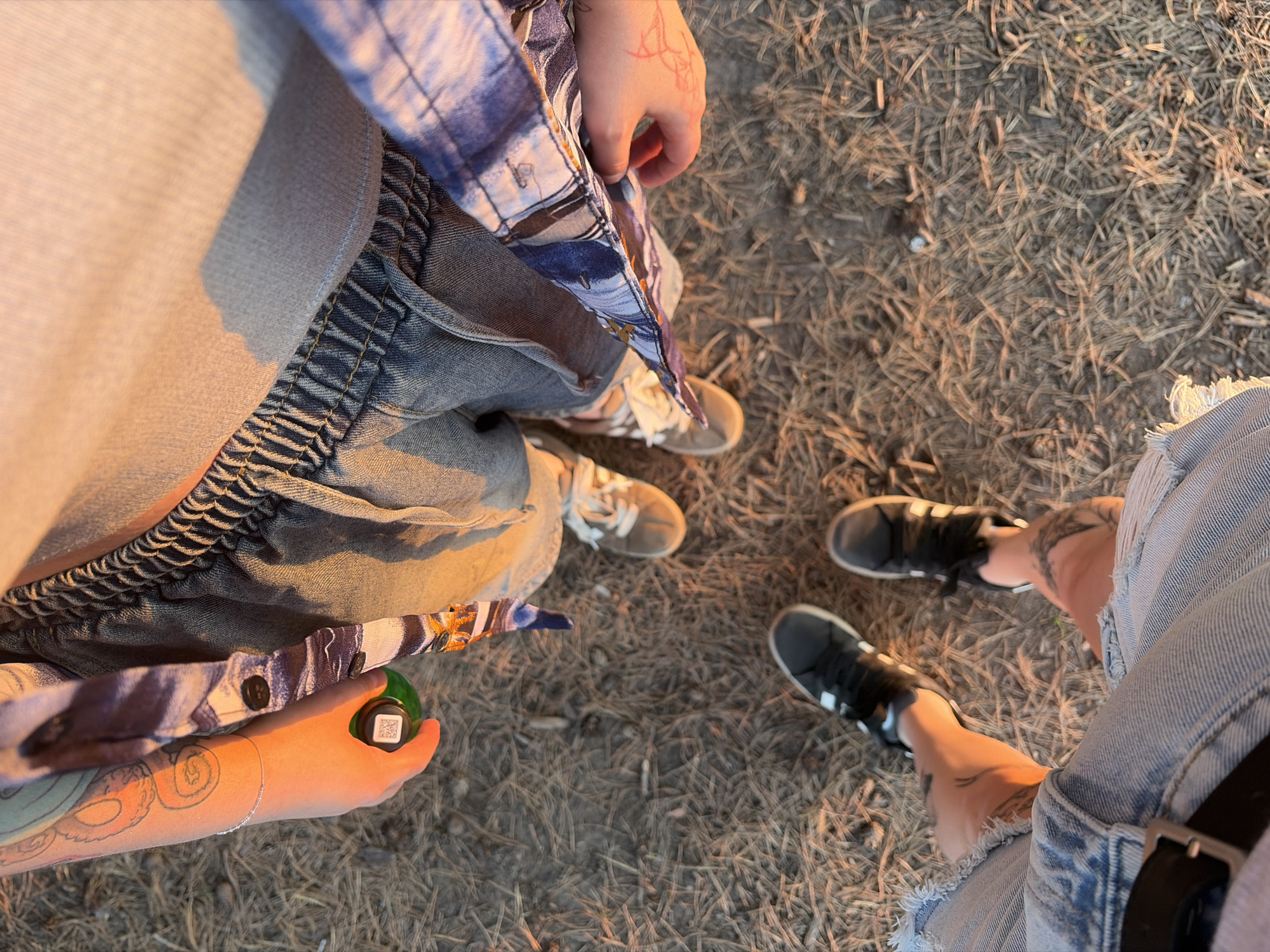 A high-angle shot shows three people standing on dry grass, possibly in a park. The person on the left is wearing light-colored sweatpants, jeans, and a partially unbuttoned blue patterned shirt. They are holding a green spherical object in their hand, which has a small white square label with a barcode on it. The person in the middle is wearing jeans and beige sneakers. The person on the right is wearing light-wash denim shorts and black shoes. The background is dry grass. No specific landmark information or city of Chaikovsky, Russia details are identifiable in the image.