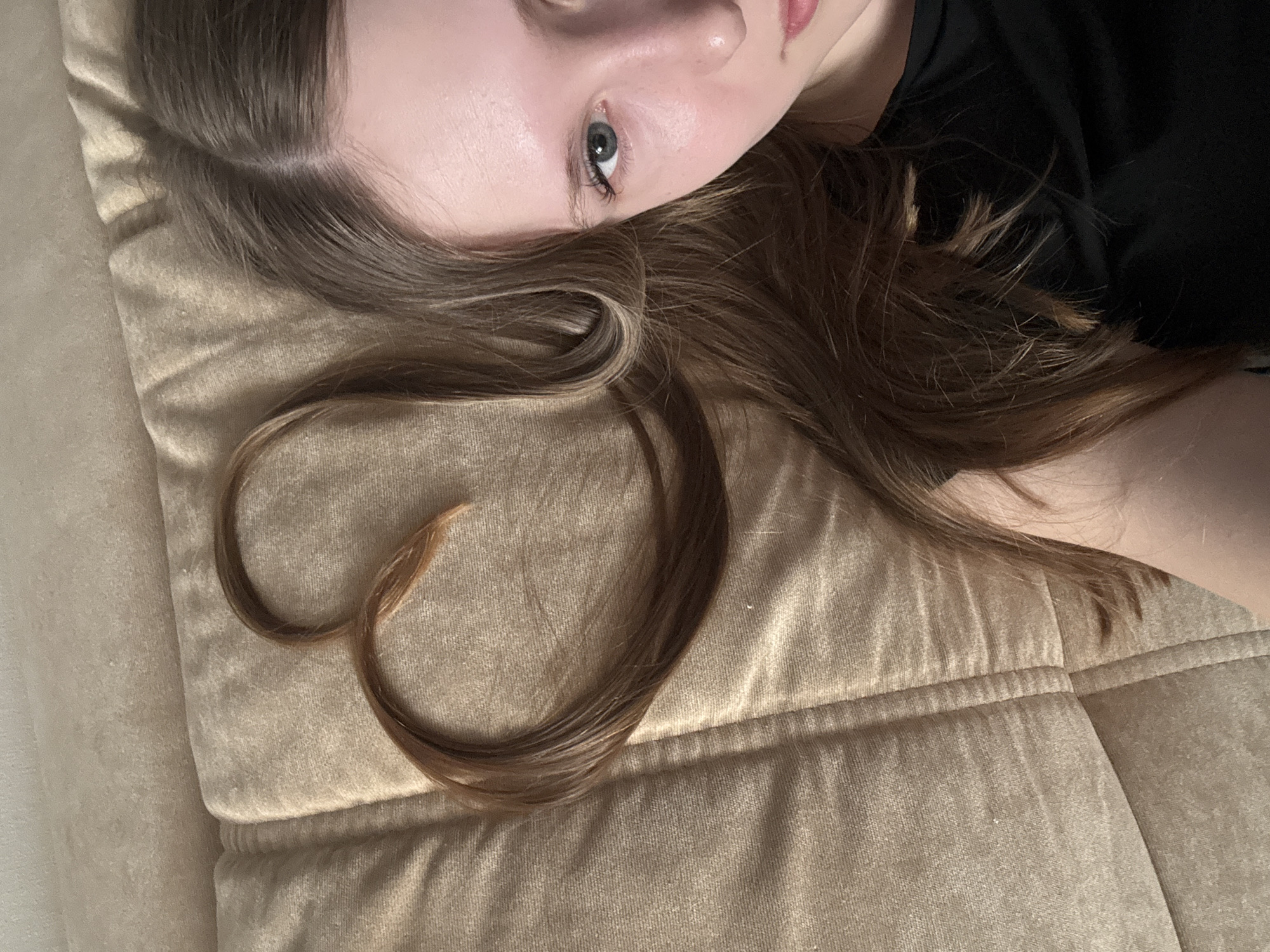 A young woman with fair skin and light brown hair lies on a tan upholstered surface, possibly a couch or bed. A strand of her hair is arranged to form a heart shape. She is wearing a dark-colored shirt. The photograph appears to be taken from an above angle. There is no information to confirm the location of this image to be in the city of Chaikovsky, Russia.