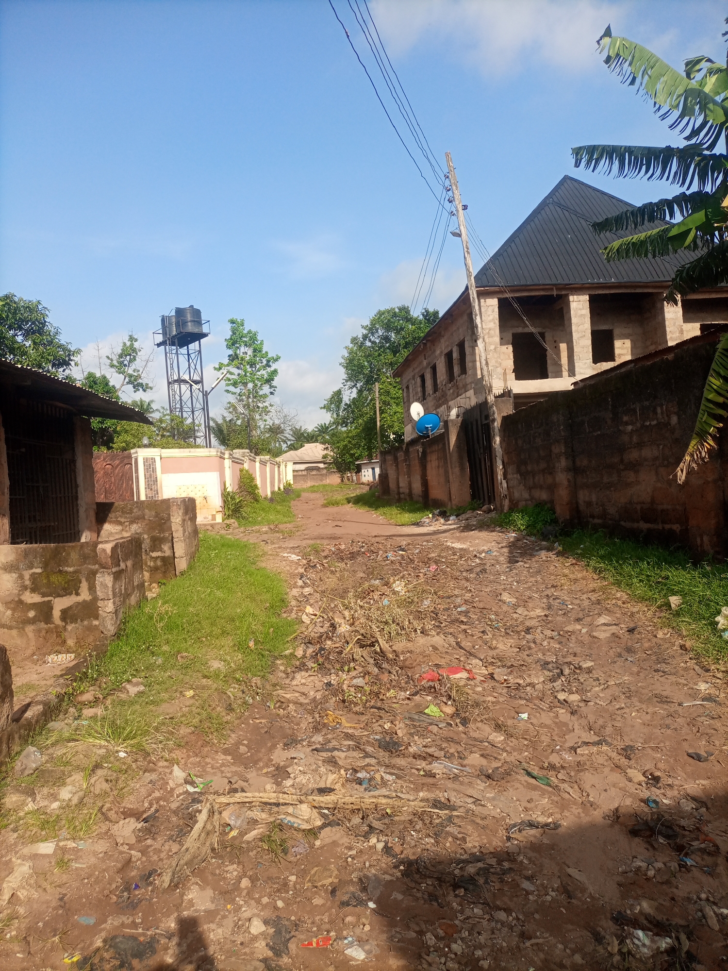 The image depicts a dusty, unpaved road in Nkpor, Nigeria. The road is lined with various structures, including a building under construction with a black metal roof on the right, and a smaller, older building with a corrugated metal roof on the left. A tall water tower stands in the background behind a fence. The ground is littered with debris, including plastic bags, bottles, and other waste. Lush green grass grows alongside the road, contrasting with the dry, barren earth. The sky is a clear blue with scattered clouds, and the sun is casting shadows, indicating it is daytime. The overall scene suggests a developing area with both new construction and signs of neglect.