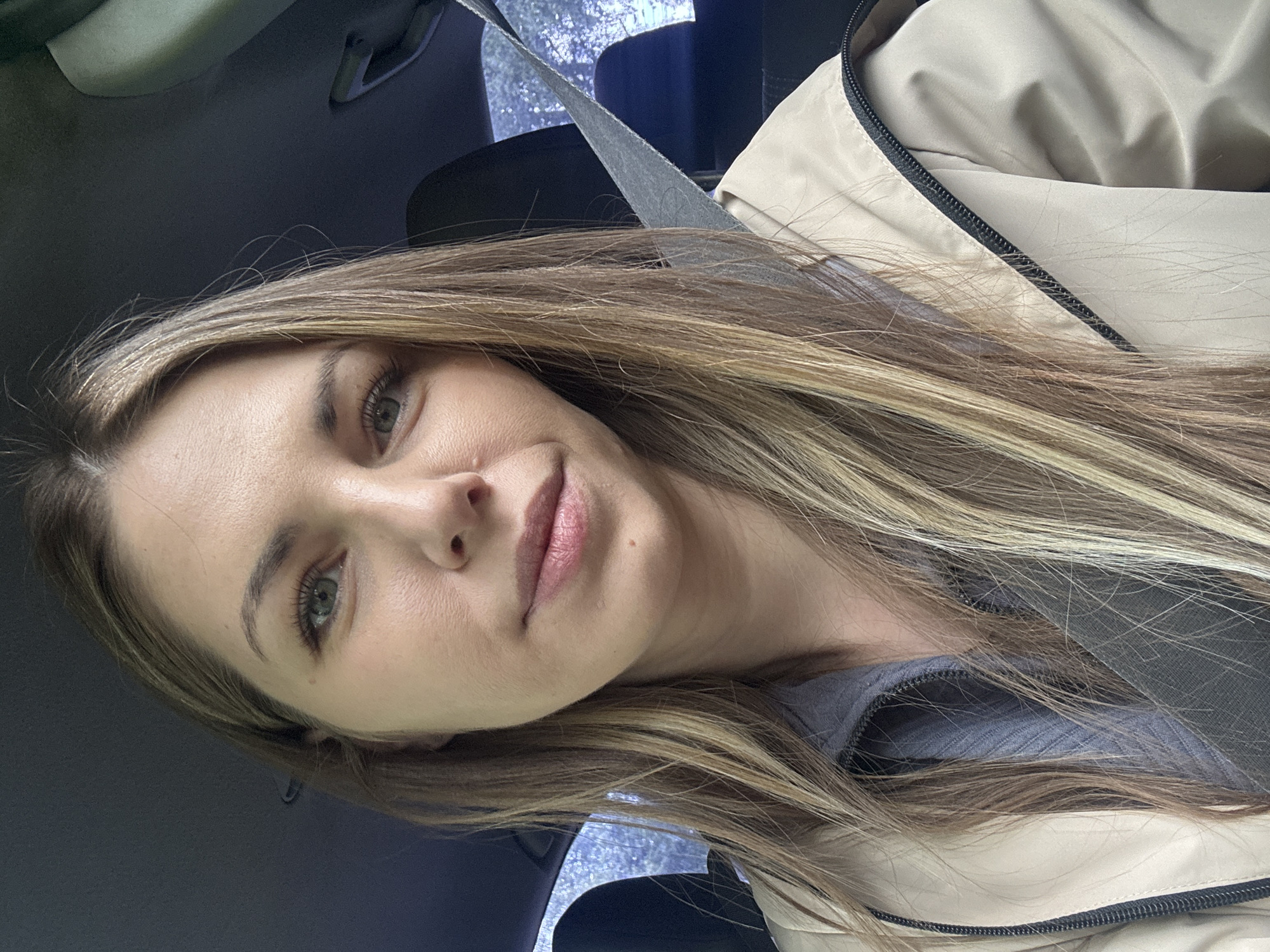 A woman with blonde and brown hair is seated in what appears to be a car. She is wearing a seatbelt and a tan-colored jacket with a black zipper. The interior of the car is visible in the background. The location is unknown.