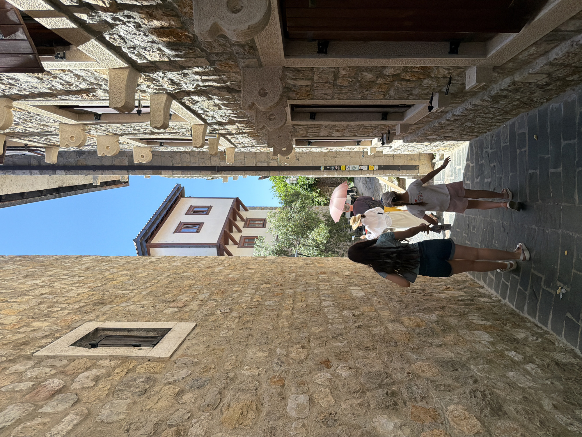 This image depicts a narrow cobblestone street in Ulcinj, Montenegro, lined with traditional stone buildings. The sun casts harsh shadows, illuminating the textured facades of the buildings. On the left, a tall, rough-hewn stone wall dominates the foreground, with a small, dark-shuttered window set into it. On the right, another stone building features multiple windows with decorative stone corbels, and a dark wooden door is visible. In the middle ground, two young girls are walking away from the camera down the street. The girl in the foreground on the left wears a turquoise top and denim shorts, while the girl on the right wears a white t-shirt and pink shorts. They are walking towards other figures further down the street, one of whom is holding a pink umbrella. In the background, a white building with red-tiled roofs and dark wooden windows is visible, suggesting an older architectural style. The sky above is a clear, bright blue. The scene captures a moment of pedestrian activity on a sunny day in a historic urban setting.