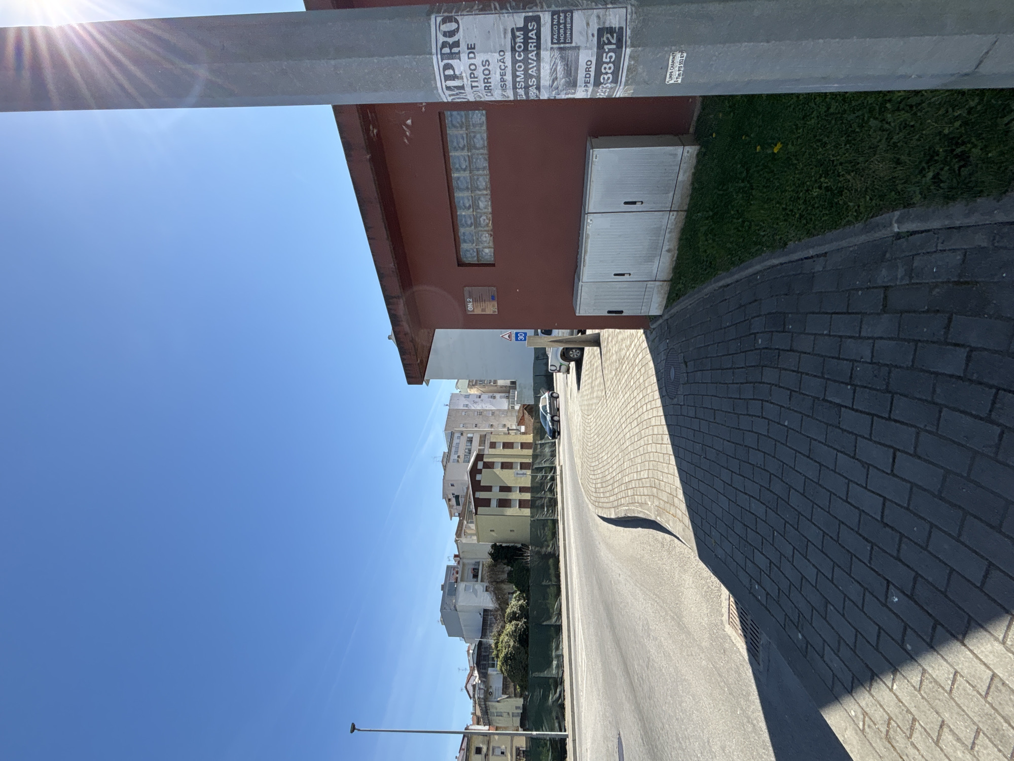 The image shows a street in Mirandela, Portugal. A red building with glass block windows and a utility box sits on the right side of the street. A sign is partially visible on the side of the building. The street is paved and has a brick-lined sidewalk. Across the street, buildings of various heights and colors are visible. A single car is parked on the street. The sky is blue. A light pole is visible on the left side of the street. A metal bar appears to be present in the upper part of the image.