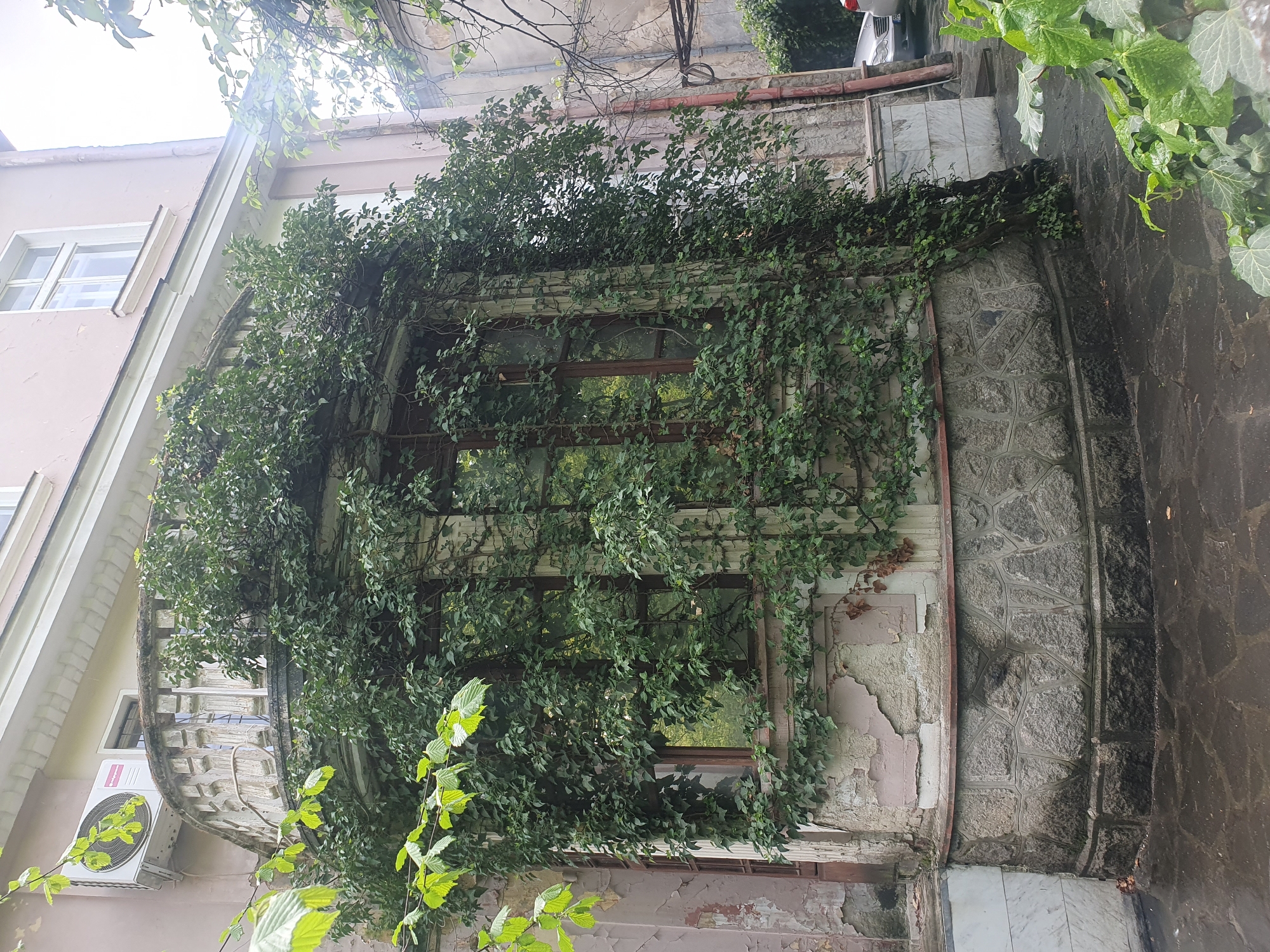An exterior shot shows a rounded bay window on a building heavily overgrown with ivy. The building has multiple windows, some with white frames. An air conditioning unit is affixed beneath the bay window. The structure of the bay window is partly visible, with cracked plaster, a dark brown wooden window frame, and a gray stone element on the right. The location is likely in the city of Plovdiv, Bulgaria, given the request.