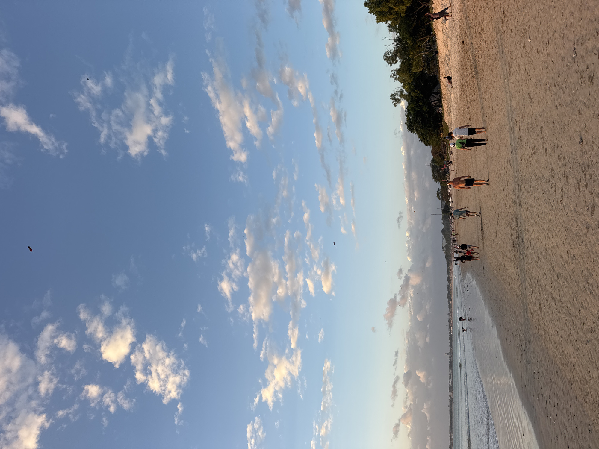 This image depicts a sunset scene at Kuta Beach in Kuta Selatan, Indonesia. The beach is populated with people walking along the shore and enjoying the evening. The sky is a clear blue, dotted with scattered white clouds, some of which are tinged with the warm hues of the setting sun. The sand is golden brown, stretching out to meet the gentle waves of the ocean. In the distance, lush green trees line the horizon, adding a natural backdrop to the scene. Several kites are visible in the sky, adding a playful element to the tranquil atmosphere.