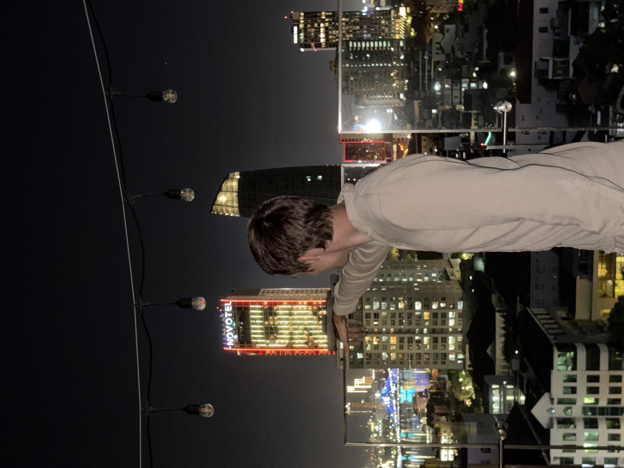A person is standing on a rooftop at night, facing away from the camera and looking out at the city skyline of Đà Nẵng, Vietnam. The person is wearing a light-colored long-sleeved shirt. In the background, the city is illuminated by the lights of numerous buildings and bridges. The Novotel Da Nang Hotel, a prominent skyscraper with red and yellow lights, is visible. String lights hang overhead, with several bulbs lit. The sky is dark, and the overall scene depicts an urban environment at night.
