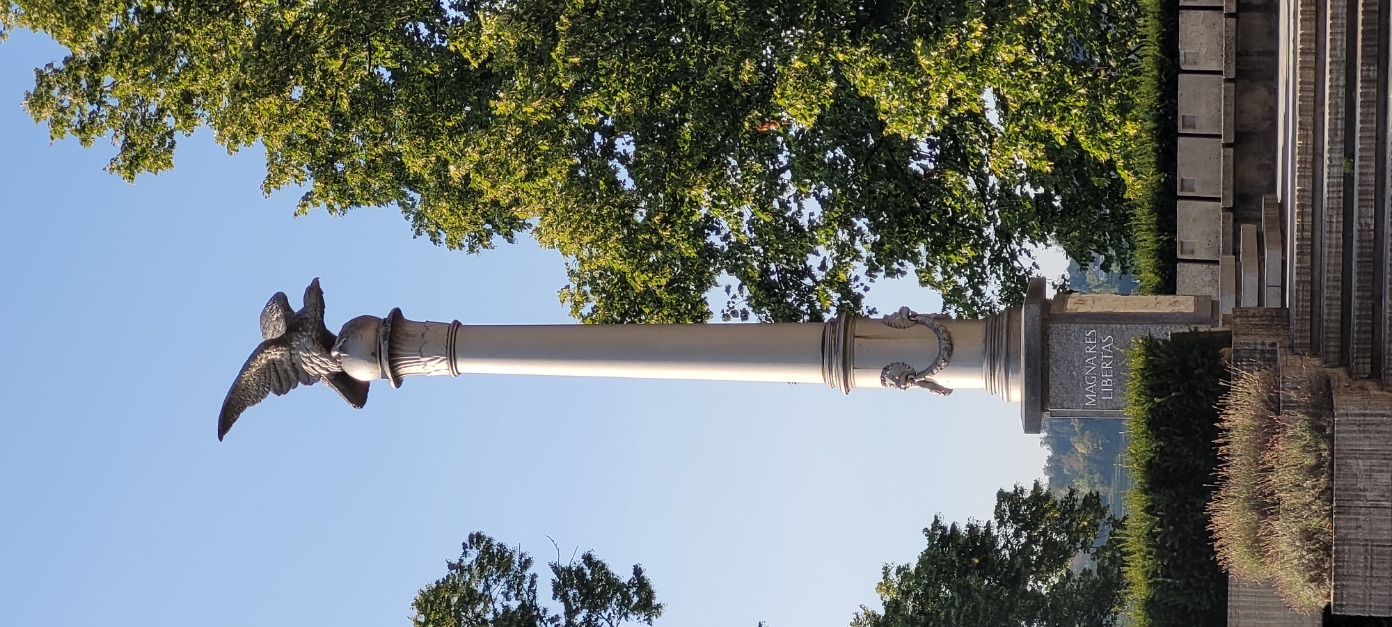 This is a vertical, eye-level, medium shot of a monument in Wattens, Austria. The monument is a tall, silver column topped with a statue of an eagle with its wings spread. The column has decorative elements, including two lion heads on either side near the base of the column. At the base of the monument, there is a granite plaque that reads "MAGNARES LIBERTAS" in white lettering. The monument is surrounded by green bushes and trees, with a clear blue sky in the background. The monument appears to be located in a park or public space, as evidenced by the stone seating area and steps in the foreground. The lighting suggests it is daytime.