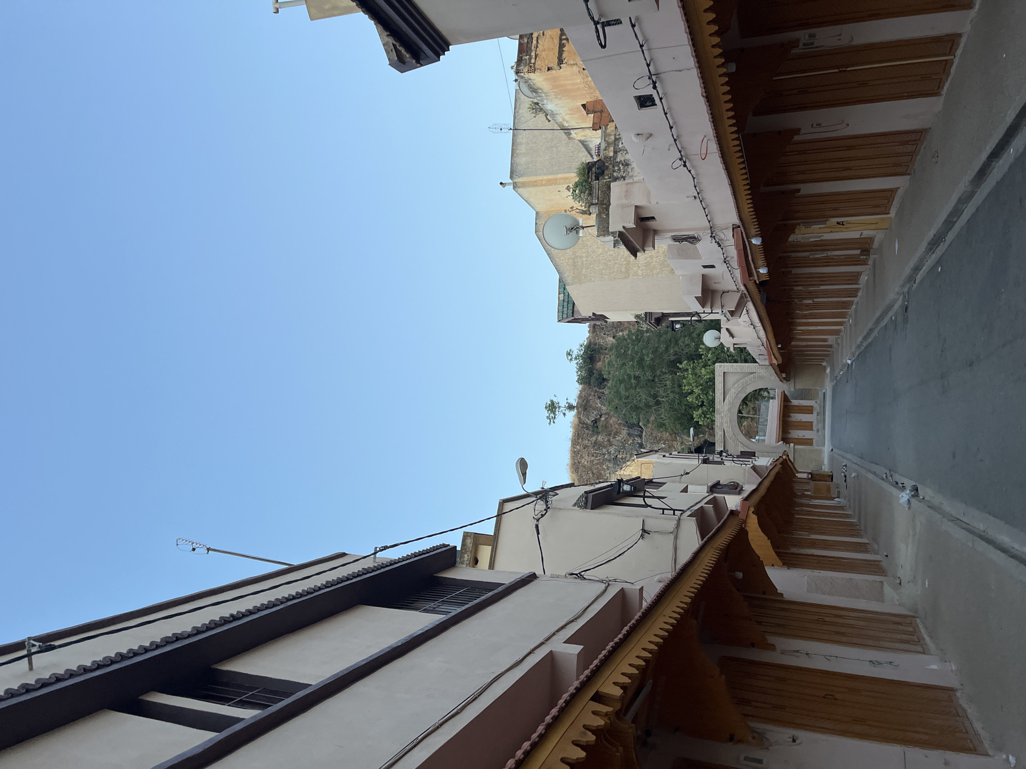 The image depicts a narrow street in El Hajeb, Morocco, lined with closed shops. The buildings are painted in neutral tones, with wooden storefronts featuring ornate awnings. The street is paved and appears to be empty of people. In the background, a rocky hill with sparse vegetation rises, and an arched gateway is visible further down the street. The sky is clear and blue.