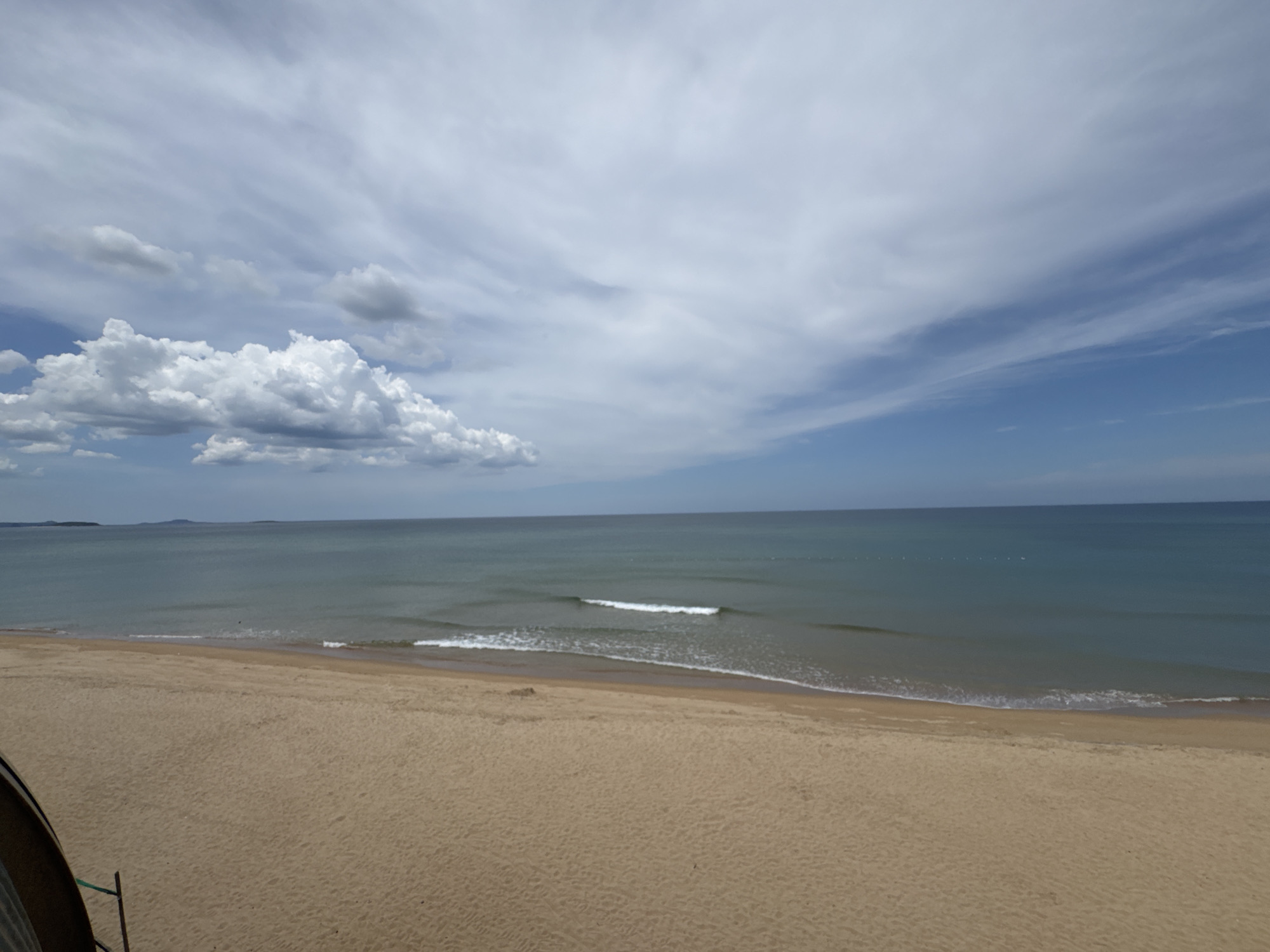 A wide, panoramic shot captures an overcast day at the beach in Tuy Hoa, Vietnam. The foreground is dominated by a vast expanse of golden sand, with some subtle indentations and a hint of a dark, curved object on the lower left. The middle ground features the gentle turquoise ocean, with a thin line of white waves breaking near the shore. In the distance, the calm sea meets a hazy horizon line, with faint outlines of landmasses visible on the far left. The sky above is a mix of light blue and scattered clouds, with some dense, white cumulus clouds clustered on the left side of the frame, and lighter, streaky cirrus clouds stretching across the upper portion. The overall impression is one of a serene, slightly moody beach scene.