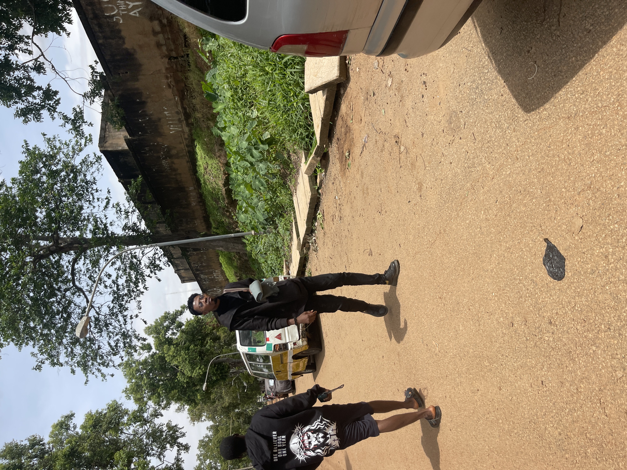 Two young men are walking on a dirt road in Rafin Par, Nigeria. The man in the foreground, on the left, is wearing a black hoodie with a graphic of Jesus Christ on the back, black shorts, and sandals. He is holding a cell phone in his left hand. The man in the background, on the right, is wearing a black turtleneck, black pants, and black shoes. He has a light-colored scarf draped over his shoulder and a dark-colored bag slung across his body.
A yellow and white van is parked on the left side of the road behind the man in the hoodie. A silver car is partially visible on the right side of the road. The road is lined with trees and a stone wall with some graffiti on it. Lush green vegetation grows along the base of the wall. The sky is overcast with some clouds. The overall environment suggests a warm, possibly humid climate.