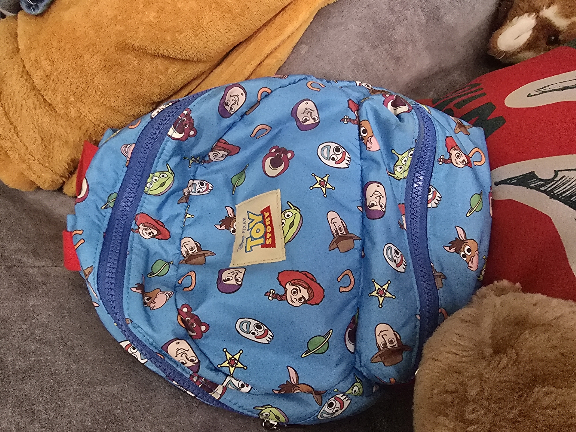 A blue backpack with a Toy Story pattern is resting on a gray couch. The backpack features various characters from the movie, including Woody, Buzz Lightyear, and aliens. The backpack has a main zippered compartment and a smaller zippered pocket on the front. A tan towel is draped over the couch behind the backpack. The image is taken from a top-down perspective.