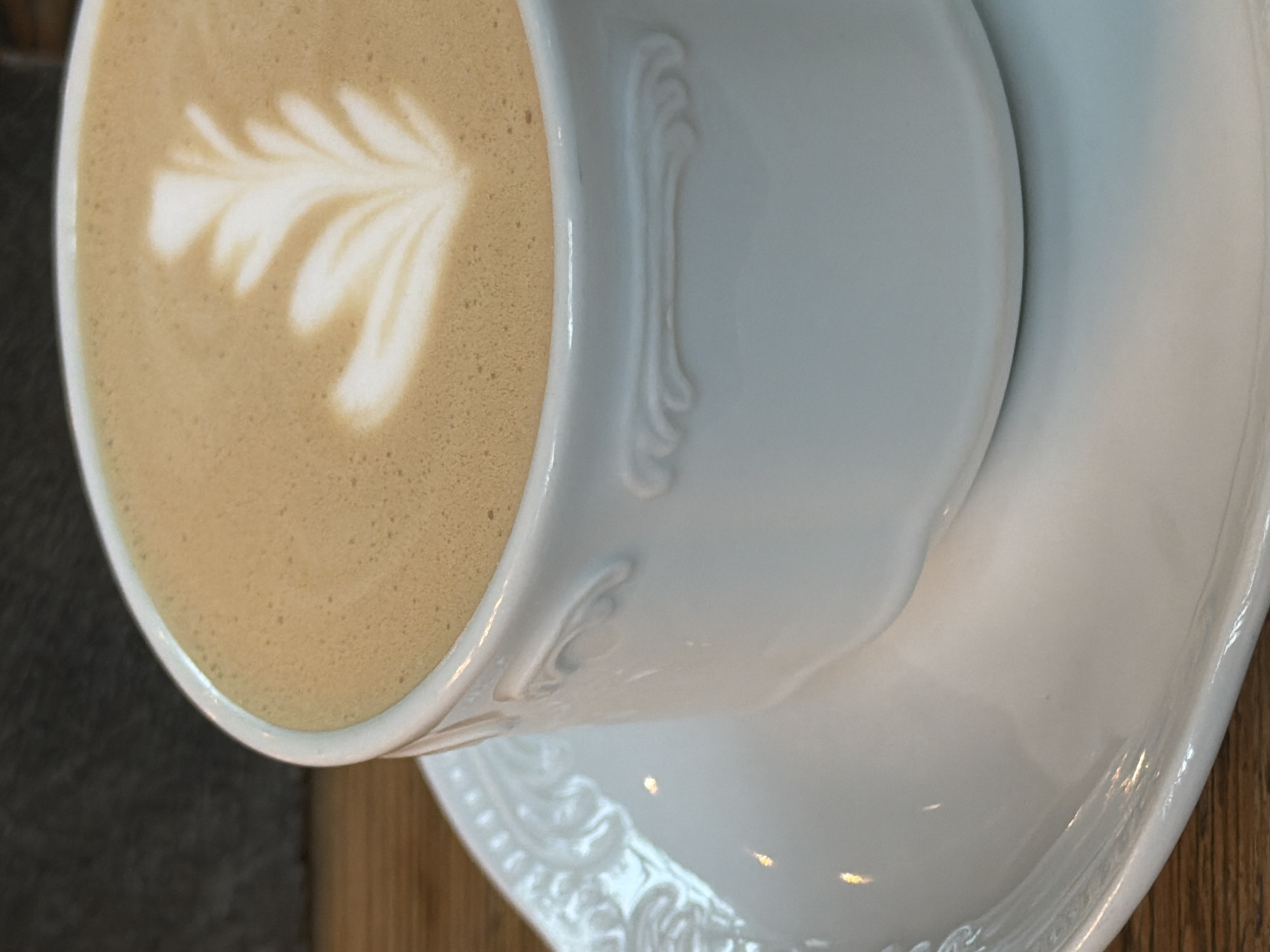 A close-up, eye-level shot shows a white ceramic coffee cup filled with coffee, topped with latte art of a leaf pattern. The cup is on a matching white ceramic saucer, which is placed on a wooden surface. The background is a dark, textured wall. The lighting is soft and highlights the foam art and the details of the cup and saucer. This image is not taken in Saint Petersburg, Russia.