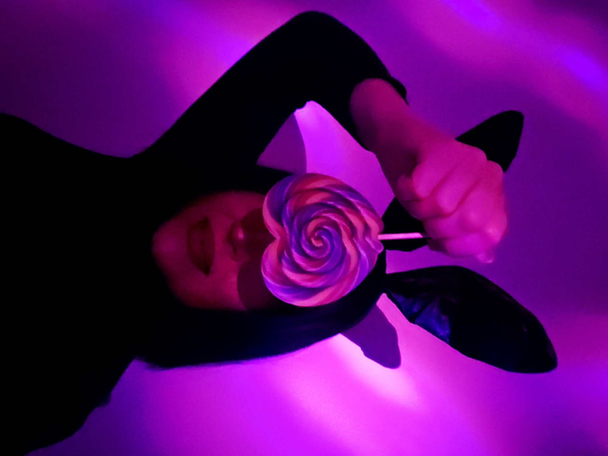 A person is lying down, wearing a black garment and bunny ears, with a heart-shaped lollipop held in front of their face. The scene is illuminated with purple lighting. The location is Unknown, Unknown.