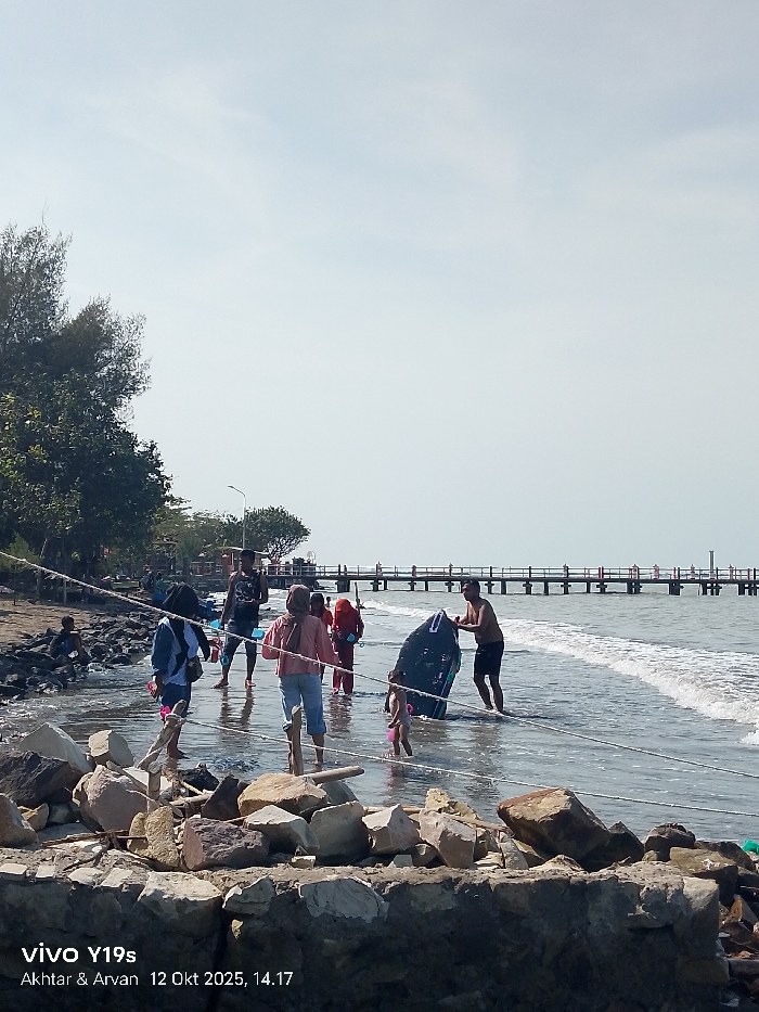 The image captures a daytime scene at a coastal location in Limbangan, Indonesia. In the foreground, a rough barrier of large, irregularly shaped rocks, primarily light brown and grey, extends horizontally across the frame. Two white ropes or lines run parallel over these rocks and into the water.
In the mid-ground, several individuals are present in the shallow ocean water. From left to right: A person wearing a light blue jacket and dark trousers stands facing away at the water's edge. Adjacent, a woman in a pink long-sleeved top, light blue jeans, and a light brown hijab wades through the water. Further right, a man in a black tank top and dark shorts stands deeper in the water. To his right, another woman in a red top and dark lower garment is also wading. On the far right, a shirtless man in dark shorts holds a dark blue boogie board or small kayak vertically in the water, positioned next to a small child in pink swimwear. One individual is seated among the rocks on the far left shore.
The ocean water shows gentle white-capped waves breaking near the shore and recedes into a darker expanse towards the horizon. In the background, a long wooden pier, supported by numerous vertical posts, extends horizontally across the water, with several indistinct figures visible walking on its surface. The sky overhead is light blue and mostly clear. On the left side of the image, dense green trees line the shore, and a street lamp post is visible among them.