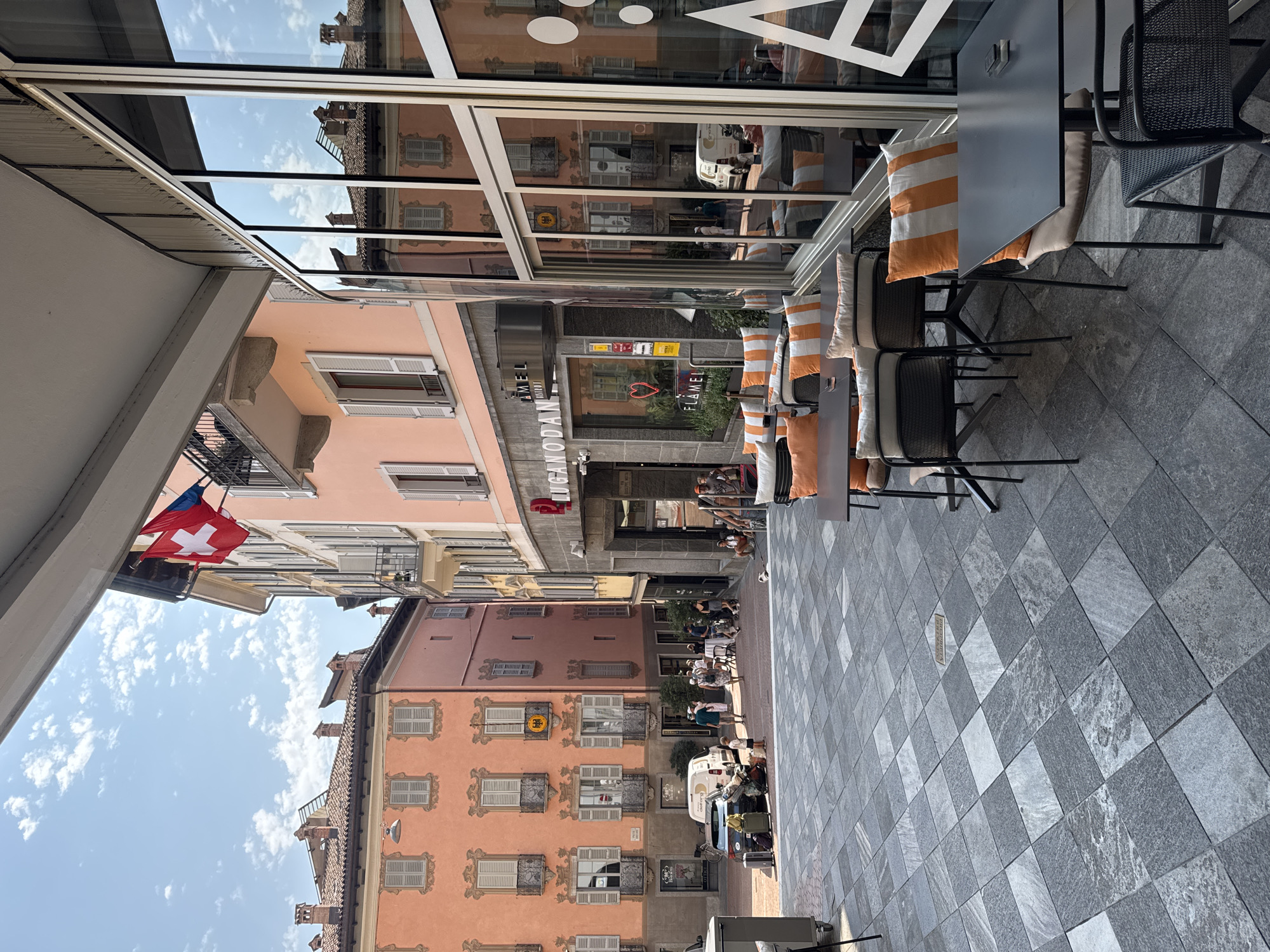 This image depicts a sunlit, European-style plaza in Lugano, Switzerland. The architecture features pastel-colored buildings with arched windows and balconies. A Swiss flag is visible flying from a balcony. In the foreground, an outdoor seating area with tables and chairs, adorned with orange and white striped cushions, suggests a cafe or restaurant. People are scattered throughout the plaza, some walking, others seated at tables. Luggage is visible near a parked car and van. The ground is paved with large, gray square tiles. The reflection of the buildings and sky is visible in the large windows of a modern establishment on the right.