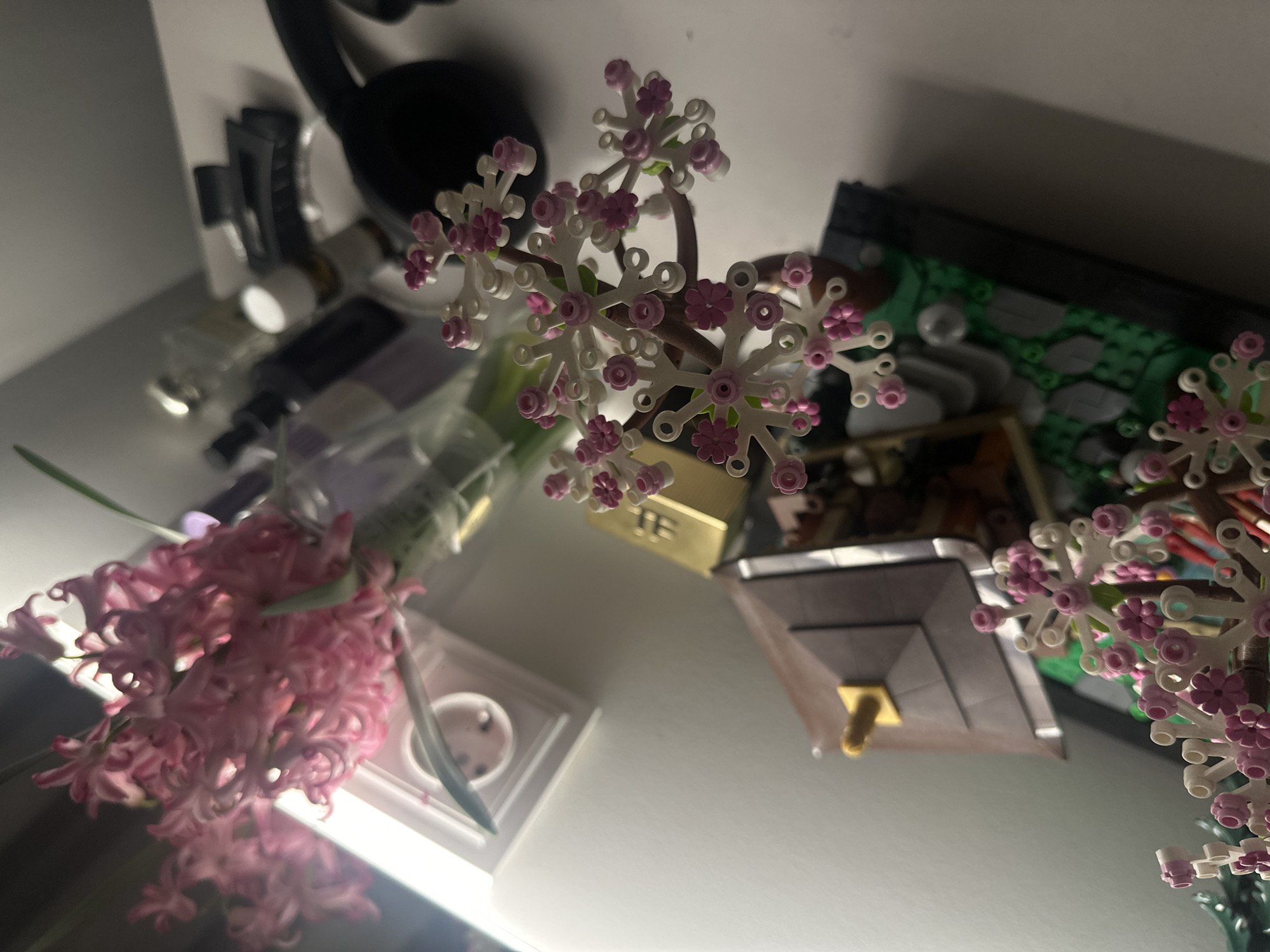 The image shows a tabletop scene. A bouquet of pink hyacinths is on the left side of the image. In the center, there is a Lego Bonsai Tree. To the right of the Bonsai Tree is a green Lego build. Other objects, including a black headset, some glass bottles, and a hair clip, are visible in the background. There is no indication that the location is Yerevan, Armenia.