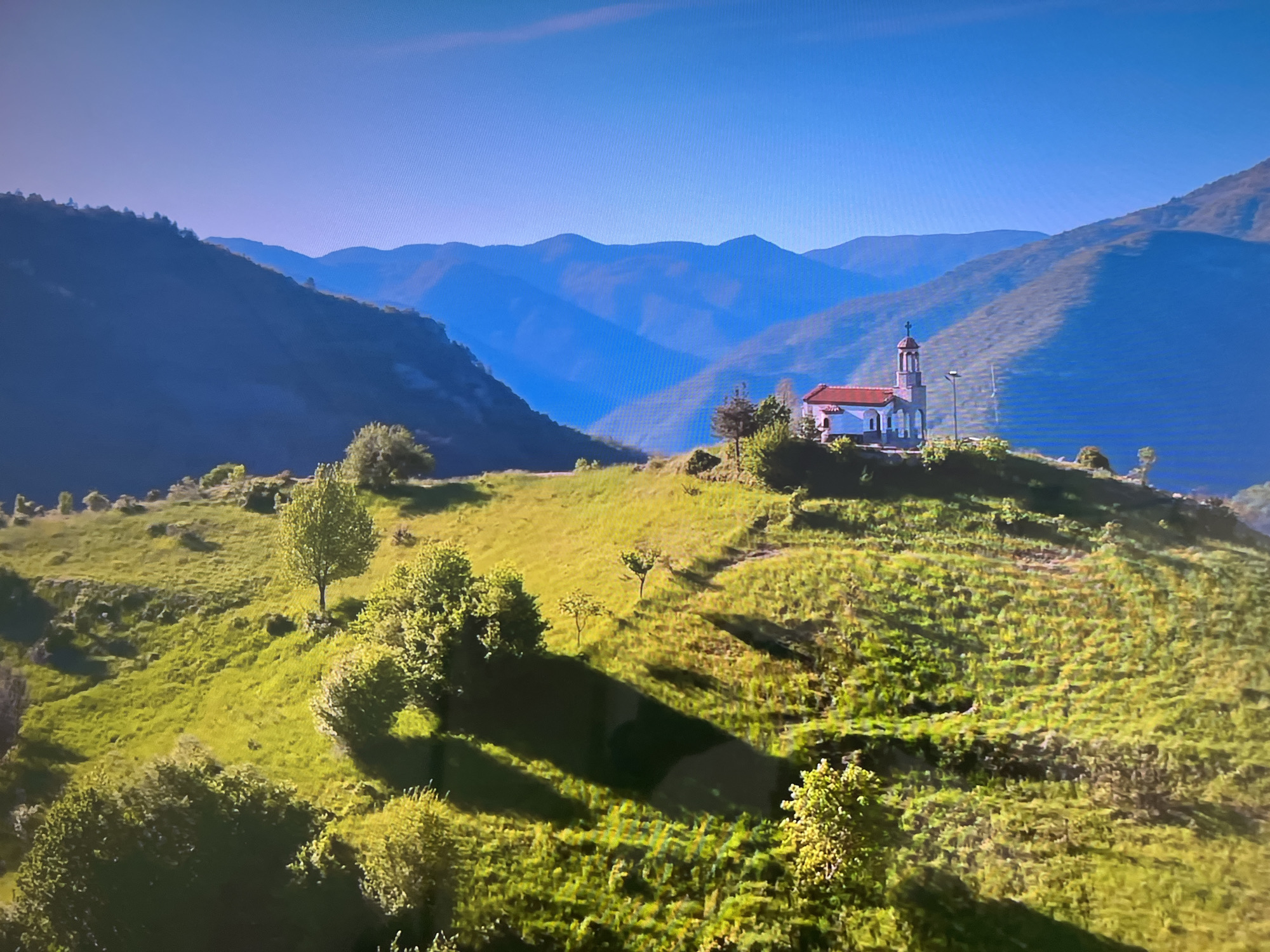 The image depicts a landscape with rolling hills and mountains in the background. A small church with a red tile roof and a steeple stands prominently on the crest of a hill. The foreground consists of green, grassy slopes with scattered trees and bushes. The mountains in the distance are layered and appear in shades of blue, indicating atmospheric perspective. The sky is clear and blue. The lighting suggests it is daytime. The location is Unknown, Unknown.