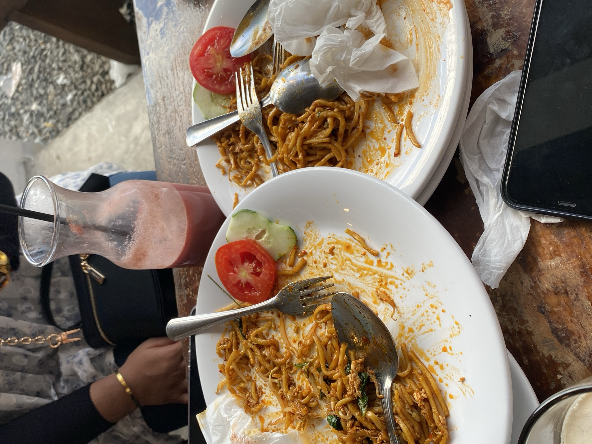 The image presents an overhead view of a dining scene, likely after a meal, in Keudekruenggeukueh, Indonesia. Two white plates are visible, each containing remnants of noodles, possibly Mie Goreng, with tomato and cucumber slices as garnishes. Silverware (forks and spoons) is scattered on the plates, suggesting that the meal has been consumed. A glass of pink liquid, possibly a watermelon juice, sits near one plate, along with a black purse. A mobile phone rests on a crumpled tissue near the plates. A person with a gold bracelet is also seen in the frame. The scene is set on a wooden table, part of which seems worn or aged. The overall impression is that of a casual dining experience.