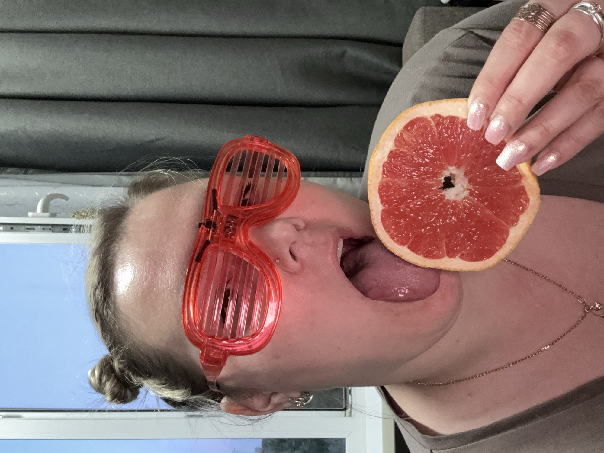A woman with red novelty glasses holds a grapefruit slice to her open mouth. She has her tongue extended, touching the fruit. The scene appears indoors, with a gray curtain in the background and a window with a view of a blue sky. The woman wears a light brown shirt and a thin necklace. Her fingernails are painted with a light pink, glittery polish, and she is wearing several rings. There is no information indicating the location is in Марковский, Russia.