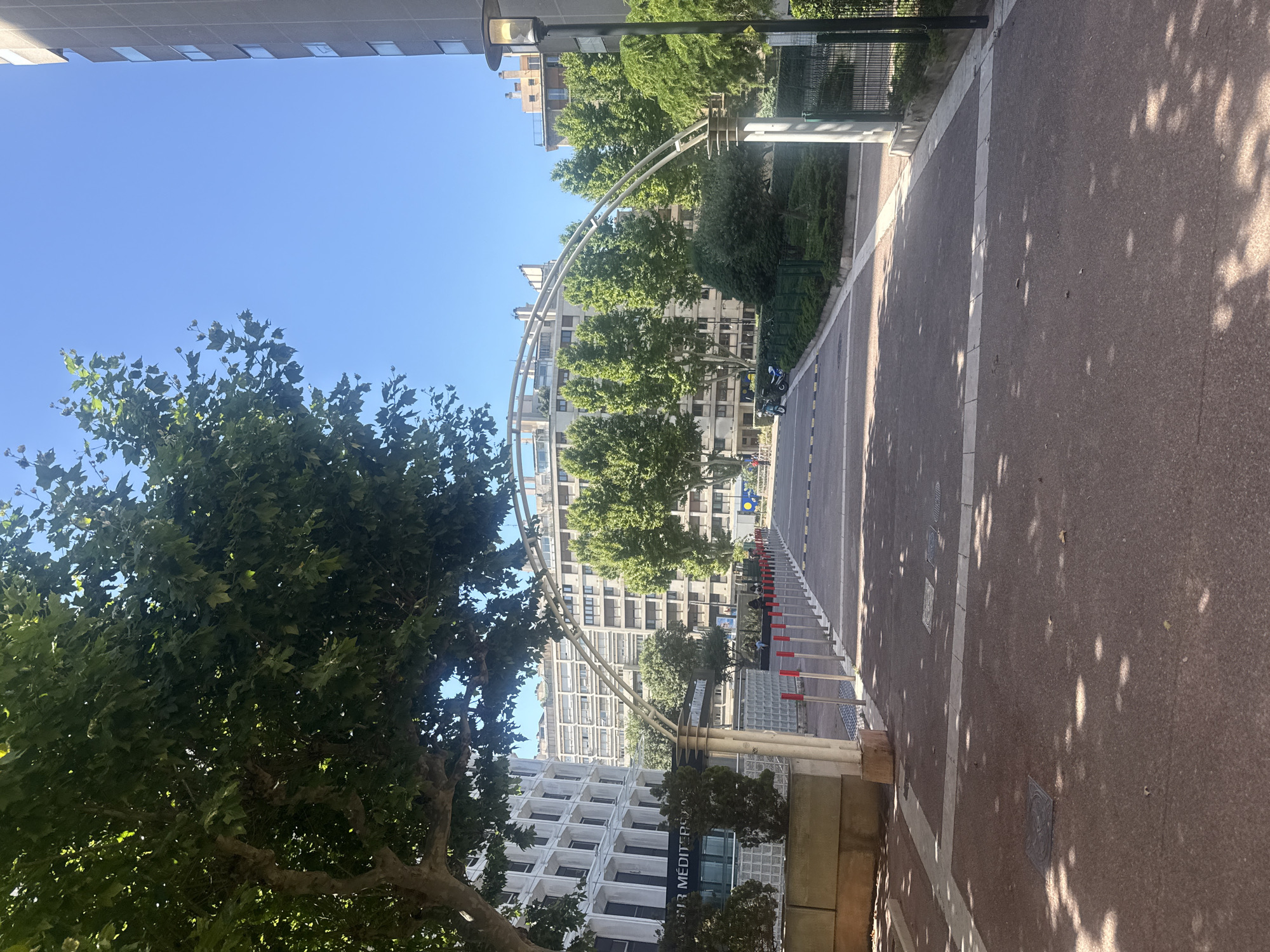 A ground-level outdoor shot shows a tree-lined pathway with a modern archway spanning across it. The pathway is paved with reddish-brown asphalt and lined with bollards. In the background, several multi-story buildings with geometric facades are visible, some with balconies and rows of windows. The sky is clear and blue. To the left, a portion of a building with the text "R MÉDITERRANÉE" is visible. Sunlight casts dappled shadows on the pathway from the trees.