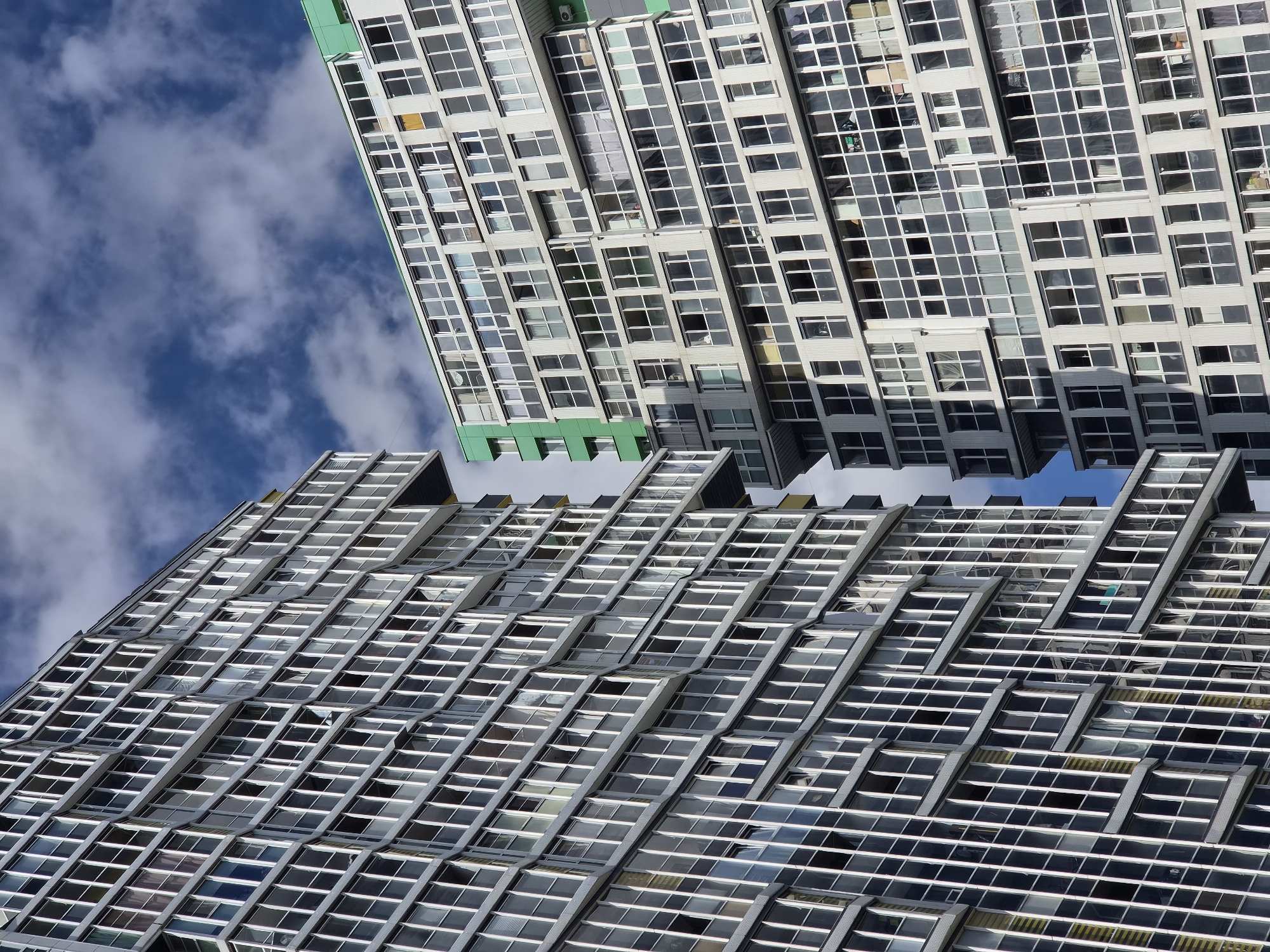 The image displays a close-up, angled view of two modern, multi-story residential buildings in Kudrovo, Russia. The buildings feature a modular design with numerous windows, creating a repetitive geometric pattern across their facades. The building on the left is characterized by a predominantly gray exterior with a multitude of tall, narrow windows, some of which appear to be open. The building on the right has a lighter-colored exterior, also with many windows, and a distinct section with a bright green facade. The sky above is a mix of blue with scattered white clouds, indicating daytime. The perspective suggests the photograph was taken from a lower vantage point, looking upwards at the structures.