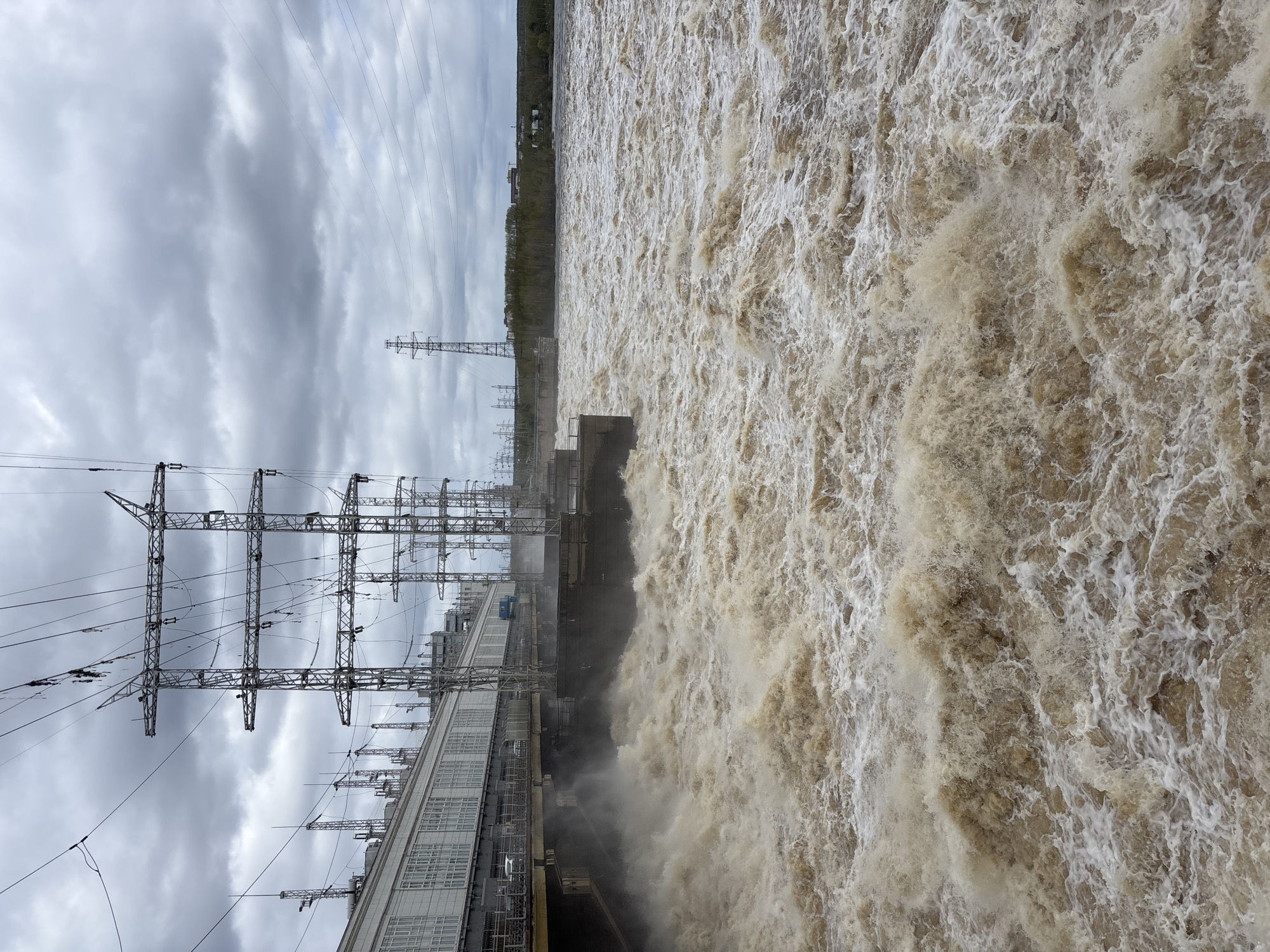 The image shows the Kama Hydroelectric Station in Perm, Russia. Turbulent, muddy water is flowing over the dam, creating a frothy surface. Power lines and transmission towers are visible on the left side of the image, near a long, light-colored building, likely part of the power station complex. The sky is overcast with gray clouds. A strip of vegetation can be seen in the background, above the dam structure.