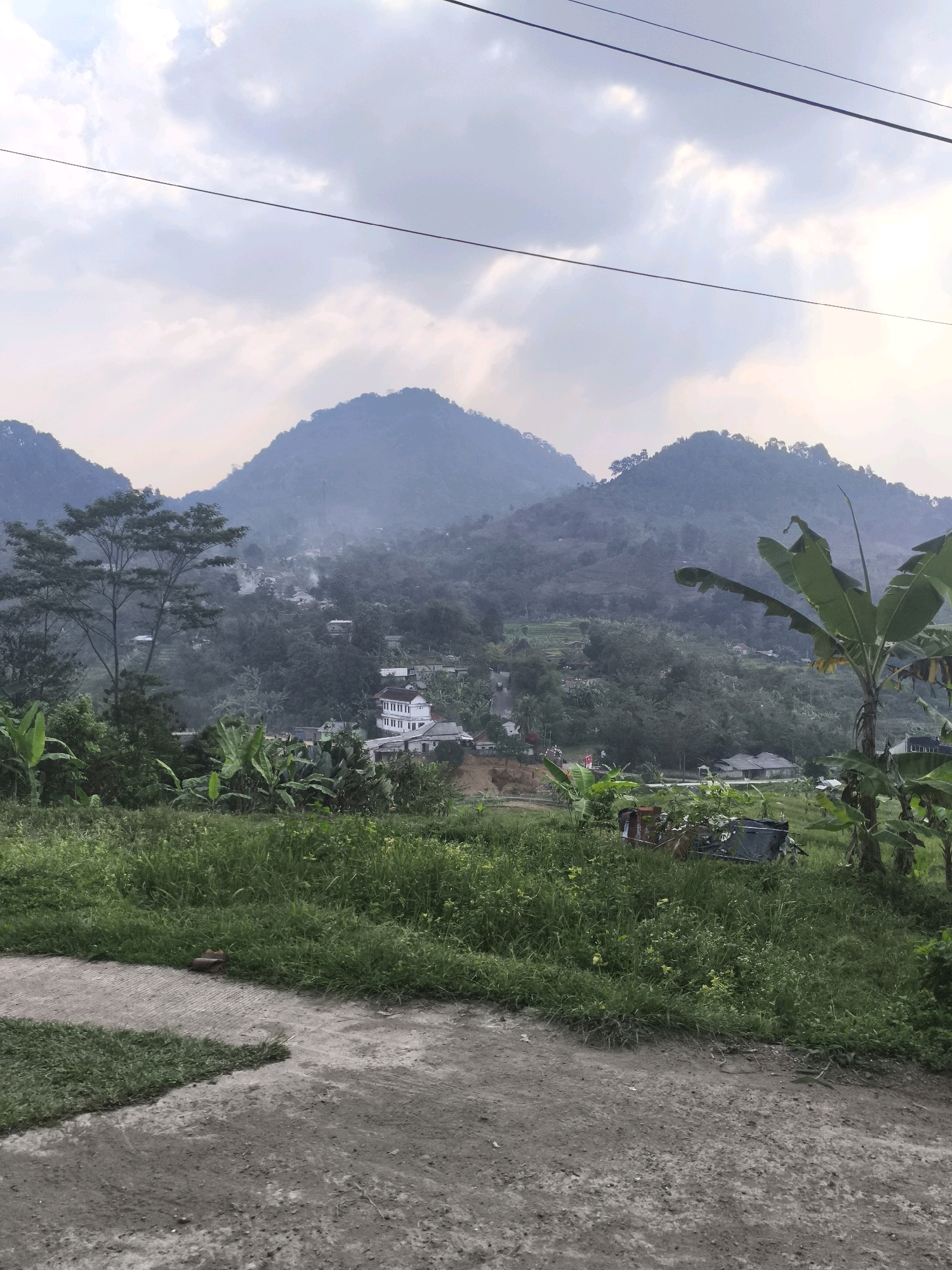 The image depicts a view of a village nestled in a valley surrounded by lush green hills and mountains. The sky is overcast with soft, diffused light. In the foreground, a dirt path leads into an area covered with tall green grass and scattered yellow wildflowers. Banana trees and other tropical vegetation are visible to the left and right.
In the middle ground, a collection of white buildings, possibly houses or a school, are clustered together in the valley. Smoke or mist rises from some areas, suggesting a humid or possibly burning environment. The buildings are surrounded by dense greenery, with some roads winding through the village.
The background is dominated by a range of mist-covered mountains that fade into the hazy sky. Power lines are visible stretching across the upper portion of the image, indicating the presence of infrastructure. The overall scene conveys a serene, rural atmosphere of a village in Indonesia.