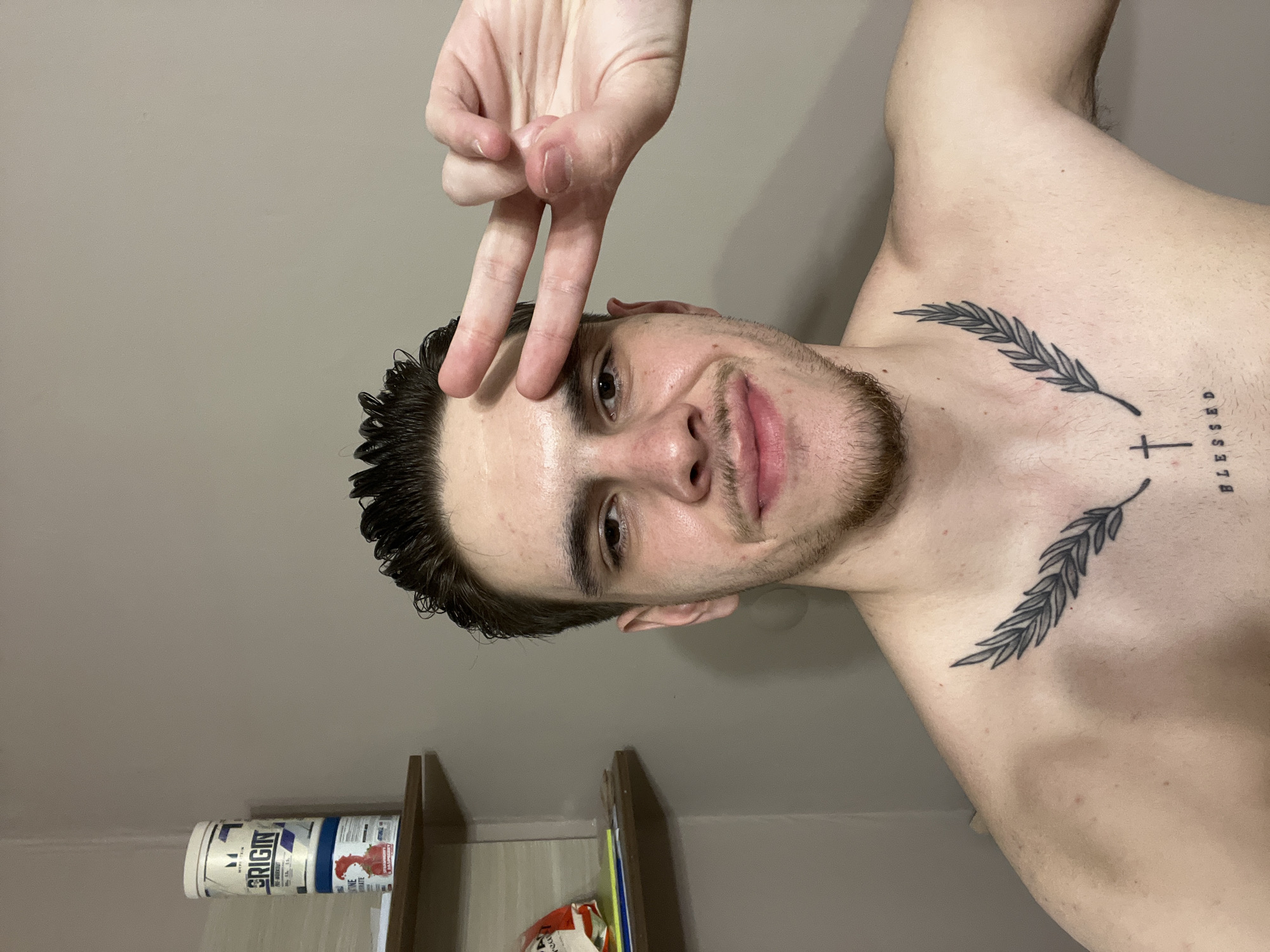 A shirtless fair-skinned male is indoors in a room with beige walls. He is positioned so that he appears horizontal in the frame. The man has dark hair, brown eyes, a trimmed beard, and is making a "peace" sign near his forehead. He has a tattoo on his chest consisting of two olive branches and the word "BLESSED" next to a cross. A shelf with a supplement container on it is visible in the background. The image provides no information regarding the city of Sofia, Bulgaria.