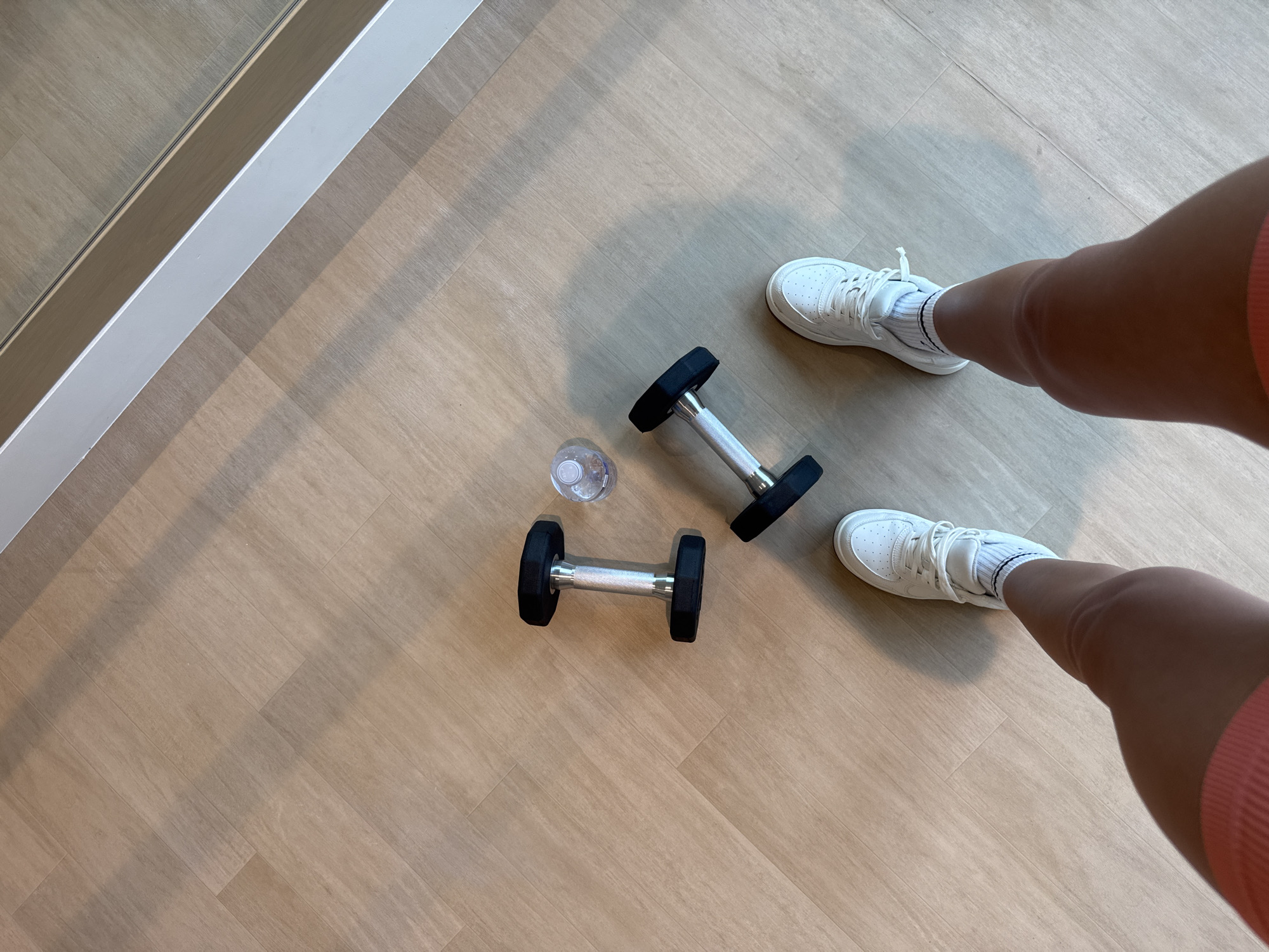 An overhead shot captures a person's legs and feet, clad in white sneakers and socks, standing on a light-colored wooden floor. Two dumbbells are laid out on the floor, one to the left and one to the right, both featuring black hexagonal ends and silver handles. Between the dumbbells, a small, clear plastic water bottle is visible. The floorboards are arranged diagonally, and a reflection of what appears to be a mirror can be seen in the upper right corner of the image. The scene is lit with soft lighting, casting gentle shadows.
