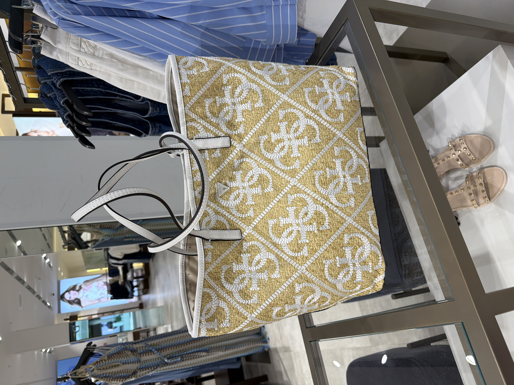 In a retail environment, a yellow and white woven tote bag with a repeating pattern of the "G" logo is displayed on a glass-topped shelf. The bag has white handles and a white trim. Behind the bag, clothing is visible on racks, including blue and white striped shirts, denim garments, and a patterned dress. A digital advertisement featuring a woman wearing a floral top is displayed on a screen in the background. A pair of light pink studded sandals are on a lower shelf. The scene appears to be in a department store or boutique.