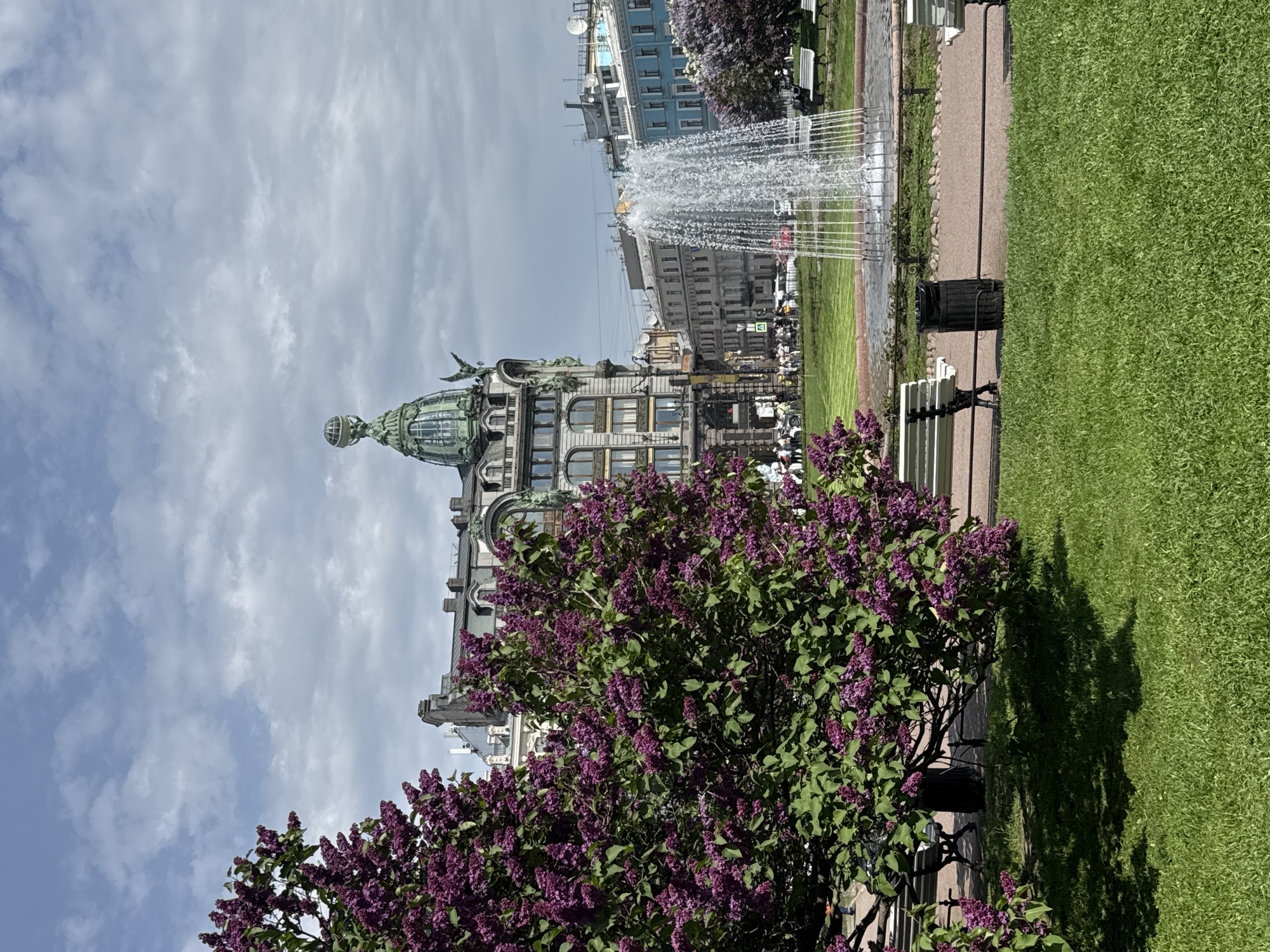 The image is a vertically-oriented shot of a cityscape on a partly cloudy day in Saint Petersburg, Russia. The Singer House, an Art Nouveau building distinguished by its corner tower and glass dome, is partially visible behind a large lilac bush with profuse purple flowers.
To the right, there is a green lawn bordered by a paved walkway with benches. A fountain is visible in the background. The sky is blue with scattered clouds. People are present in the background.