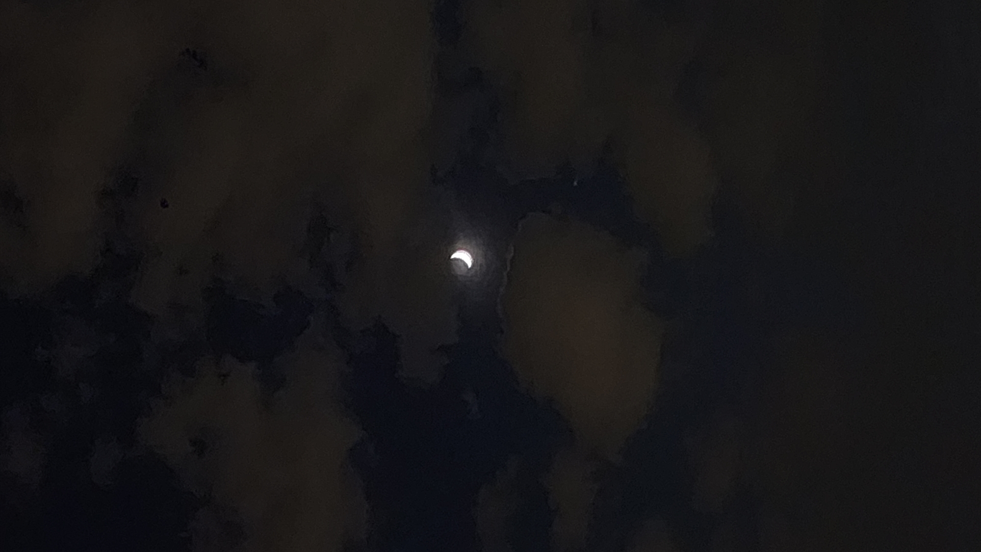 The image shows a crescent moon in a dark sky, partially obscured by clouds. The location in Moscow, Russia, is not verifiable from the image. No people or interactions are visible. The environment consists of a night sky with cloud cover and the moon.