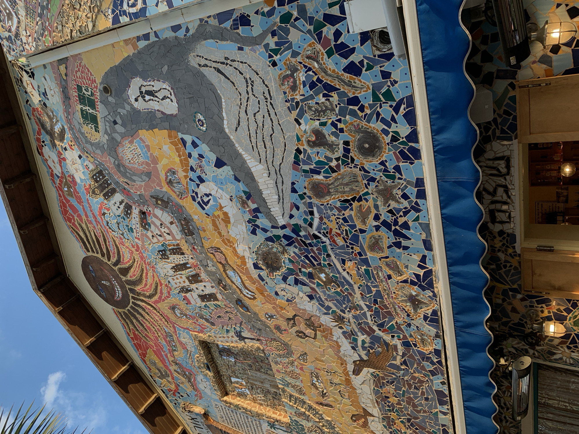 An exterior view of the historic Ilana Goor Museum's Mosaic House, located in Jaffa, Tel-Aviv, Israel, features a multi-story building facade entirely covered by an intricate, multicolored mosaic mural. The mosaic is composed of irregularly shaped ceramic and glass tesserae in various hues of blue, yellow, orange, red, brown, gray, and green.
The upper section of the mural depicts a large, smiling, sun-faced octopus or kraken-like figure with radiating tentacles extending across the facade. Below this, a cityscape with stylized buildings is rendered. To the right, a prominent gray whale-like creature is depicted, encompassing a smaller, dark stick figure within its body. The lower sections of the mosaic transition into a vibrant depiction of marine life and abstract aquatic patterns, including various fish, mollusks, and other sea creatures. A small, unglazed window opening is integrated into the mosaic on the left side, with visible Hebrew text above and to its left.
The building exhibits a pitched roof with exposed wooden eaves. Below the main mosaic, a functional blue awning with a scalloped white trim is partially retracted. Beneath the awning, additional mosaic work is visible, along with an outdoor heater and a wall-mounted lantern-style light fixture. A palm frond is visible in the upper left corner, and the sky above is clear blue with sparse white clouds.