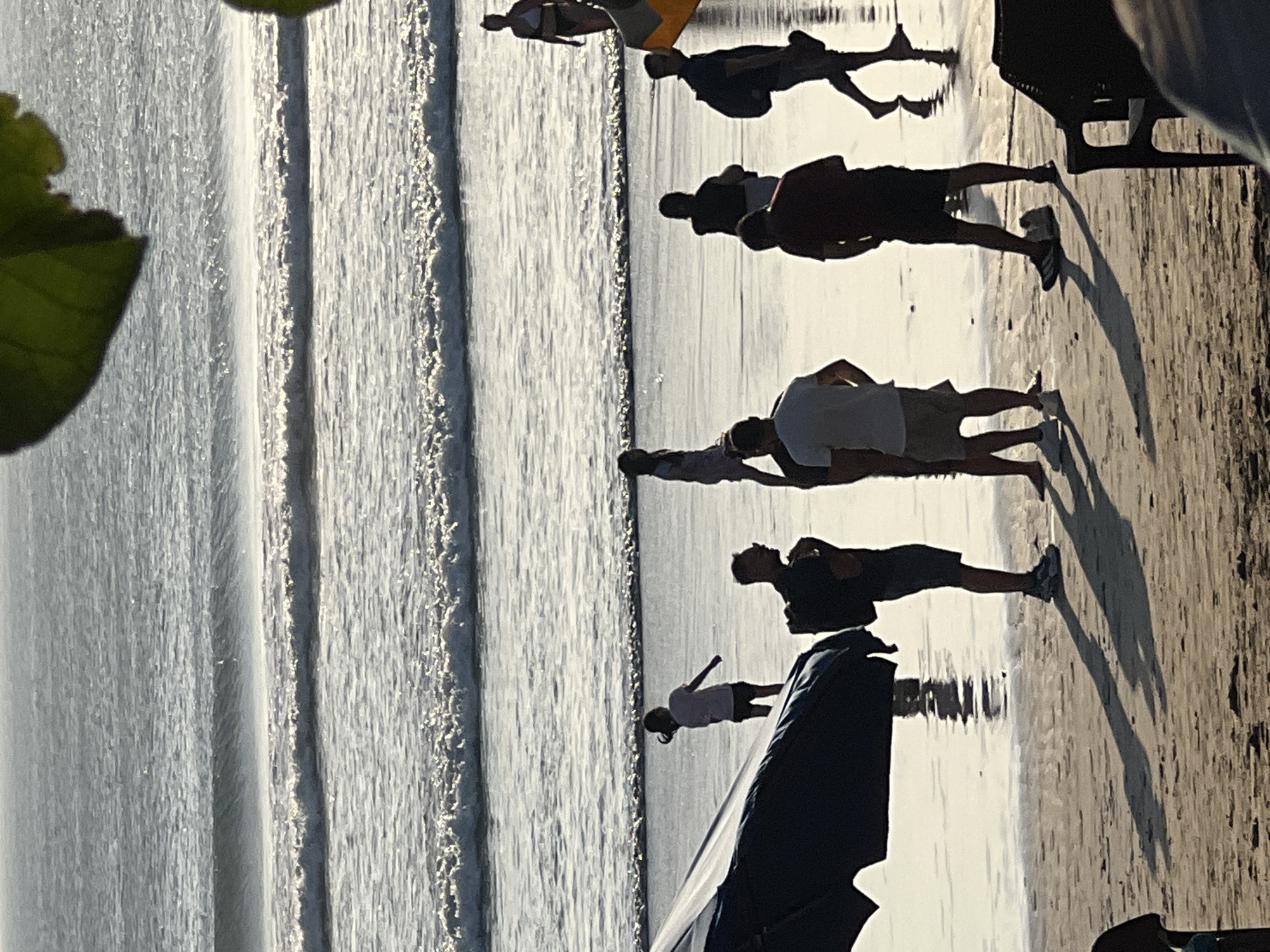 The image depicts a scene at Kuta Beach, Indonesia, during what appears to be late afternoon or early evening, given the long shadows cast by the individuals on the wet sand. The ocean is in the background, with several rows of waves breaking, creating a textured pattern of white foam against the darker water. The sky is a muted gray, indicating overcast conditions or the approach of sunset.
In the foreground, a group of people are scattered across the beach. To the left, a dark blue umbrella or canopy is partially visible, casting a large shadow. Several individuals are standing and walking on the wet sand near the water's edge. One person is wearing a white t-shirt and dark shorts, and another is in a dark t-shirt and shorts. A person in a dark t-shirt and white shorts is walking away from the camera. Further back, several other figures are visible as silhouettes against the water. One person, a child, is wearing a white t-shirt and dark shorts and is facing away from the camera, looking towards the ocean. Another individual is wearing a white top and dark bikini bottoms and is standing near the water. A person with a backpack is walking towards the right of the frame, wearing a dark t-shirt and shorts. The reflections of the people and objects are visible in the shallow water covering the sand, adding to the atmosphere of the scene. The overall mood is one of casual leisure, with people enjoying the beach.